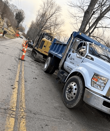 A light-colored dump truck and a yellow excavator parked on the side of a road marked by orange construction cones.