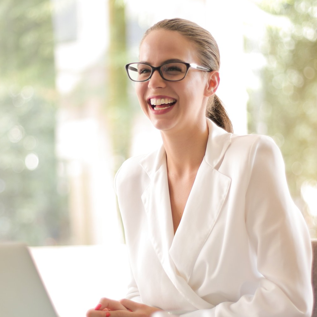 A woman wearing glasses and a white jacket is smiling in front of a laptop