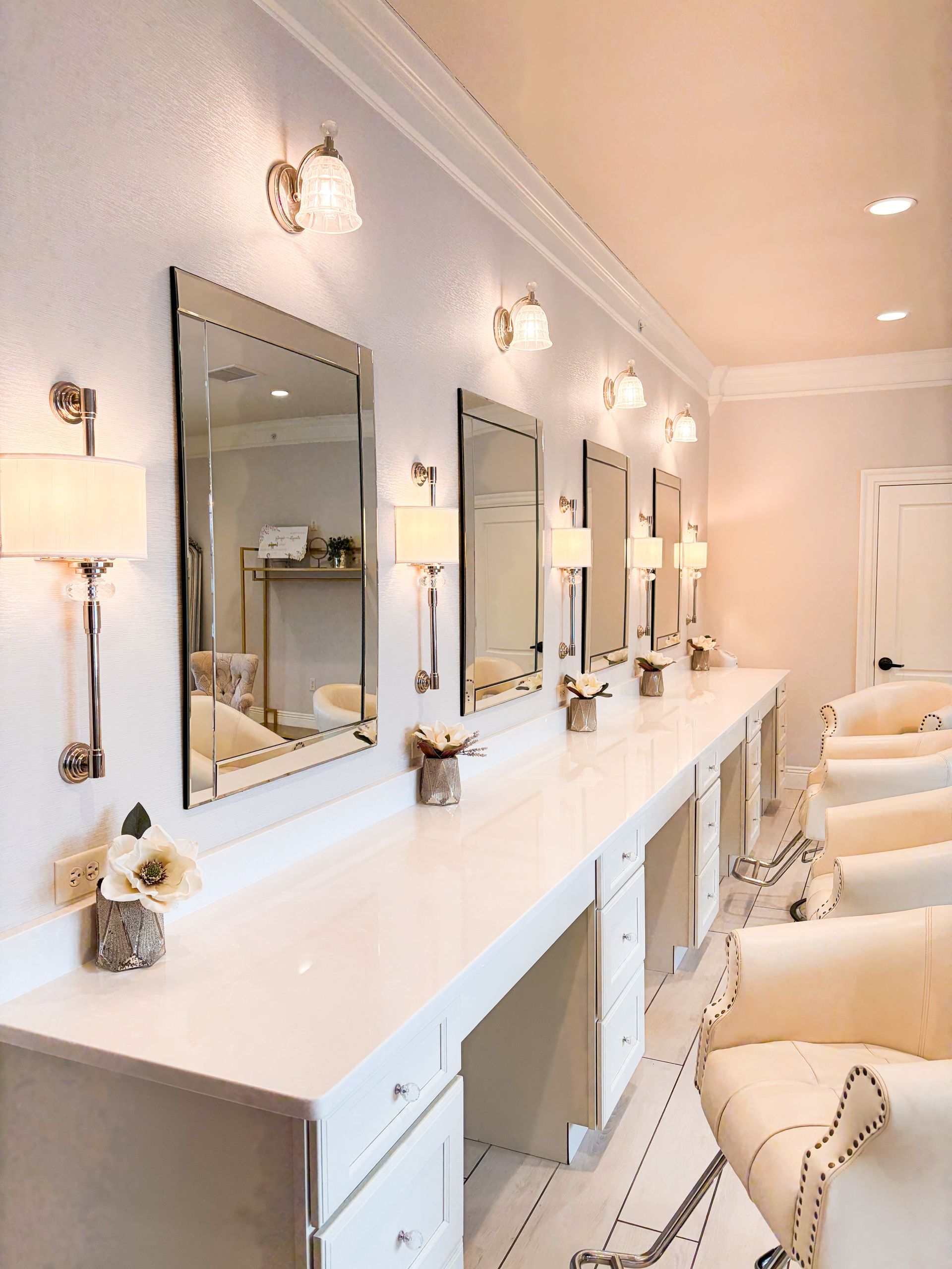 A row of white salon stations with mirrors and chairs under wall-mounted lights.