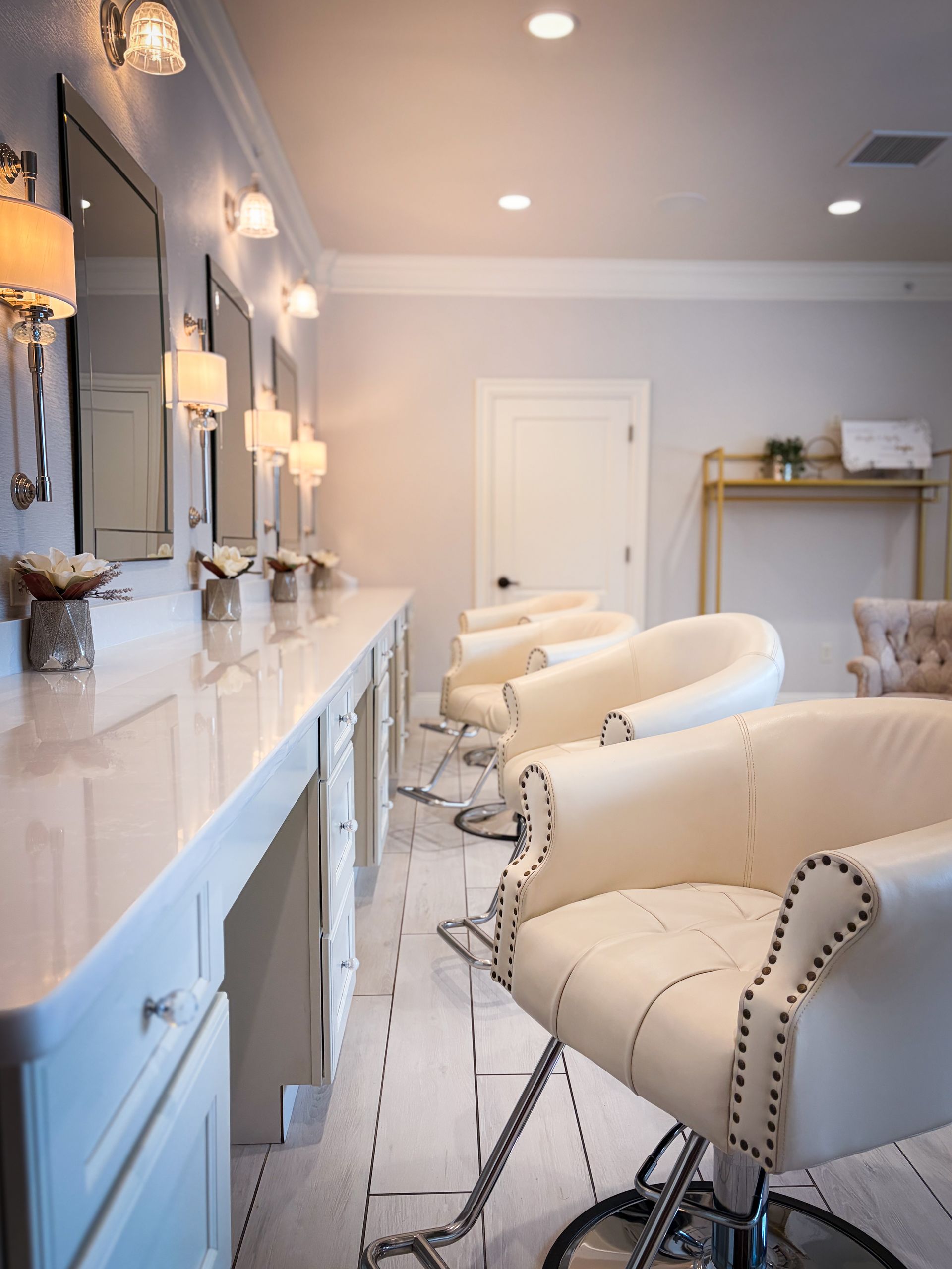 Salon with white chairs, mirrors, and lighting, next to a long counter and tiled floor.