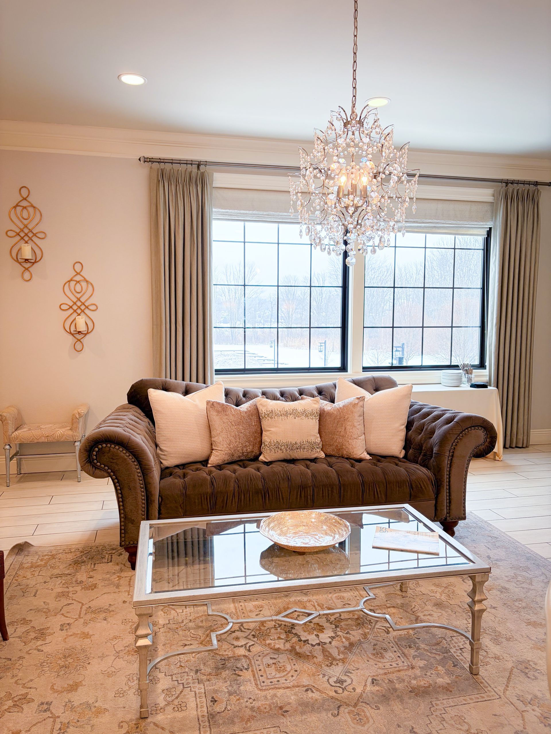 Brown tufted sofa with pink pillows, mirrored coffee table, chandelier, and window with light curtains.
