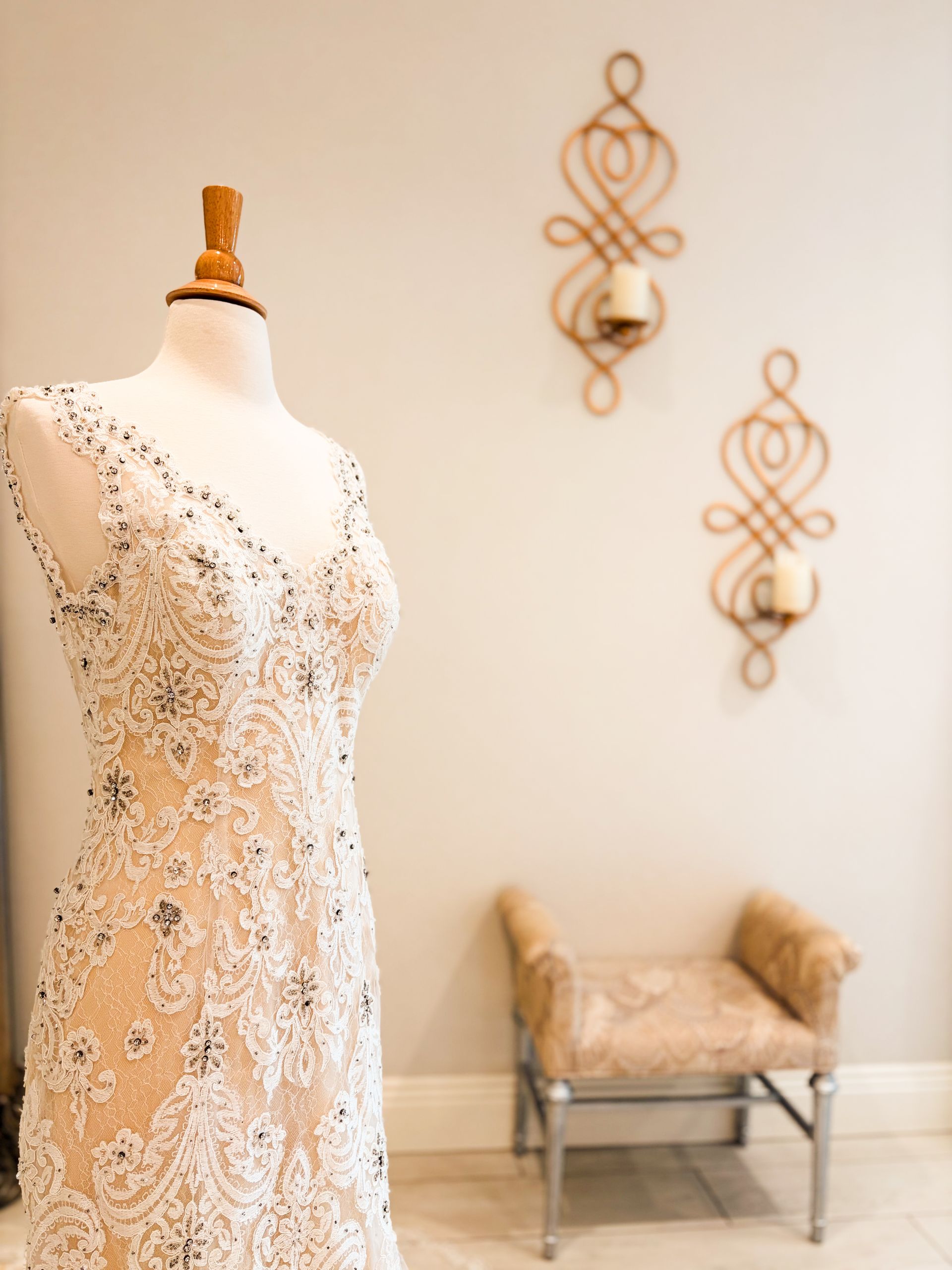 Wedding dress on a mannequin in a shop; beige and white. Decorative wall sconces and a small bench in background.