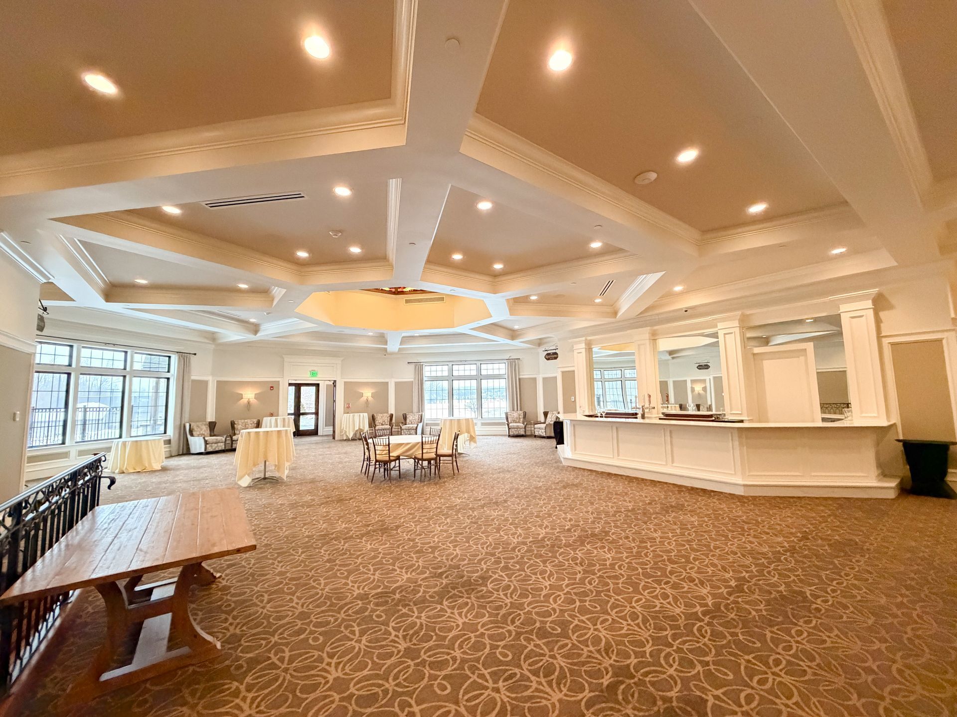 Spacious ballroom with a bar, tables, chairs, and large windows. Beige and white tones, warm lighting.