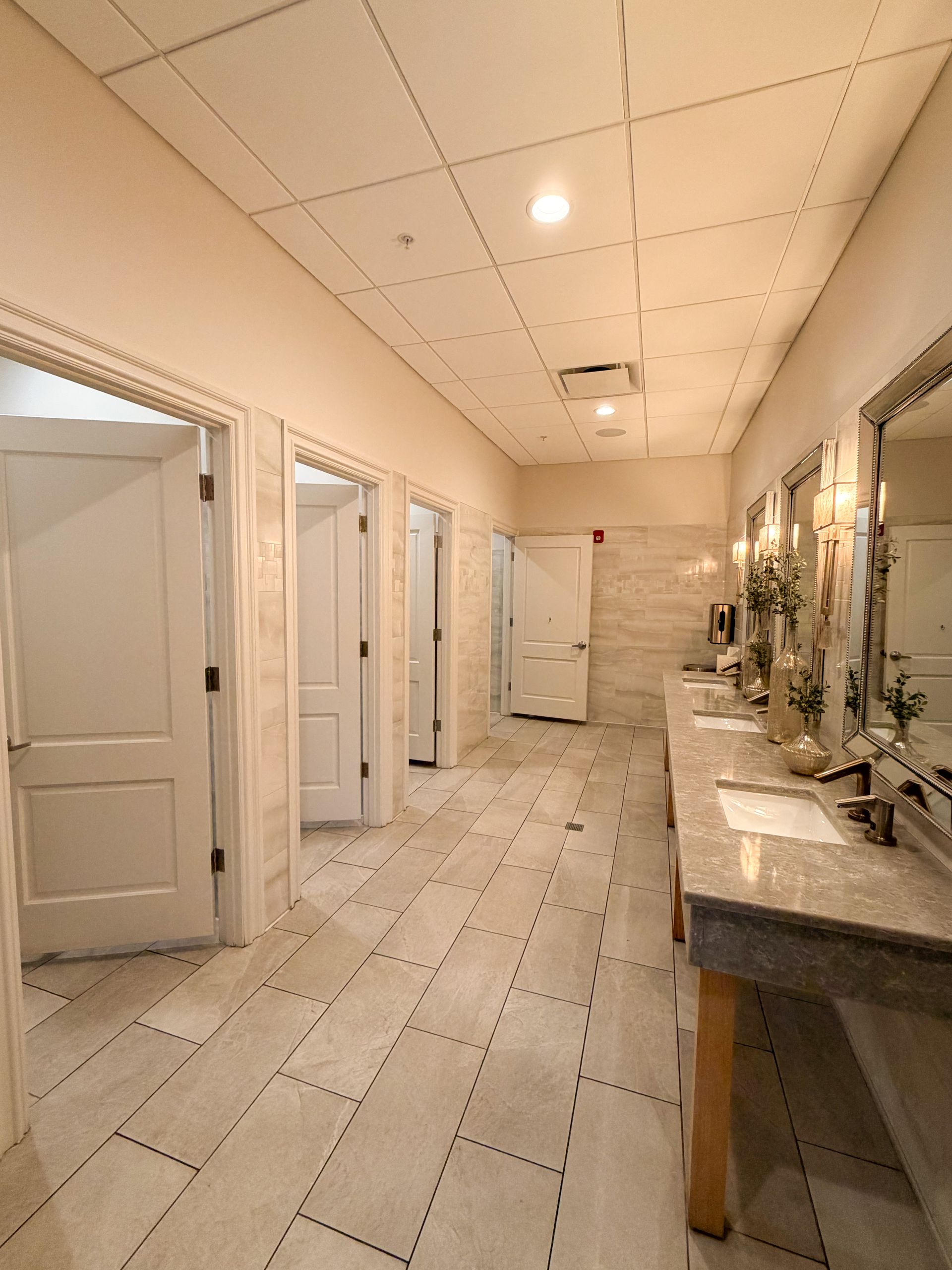 Public restroom with stalls and vanity. White doors, neutral walls, marble-like floor.
