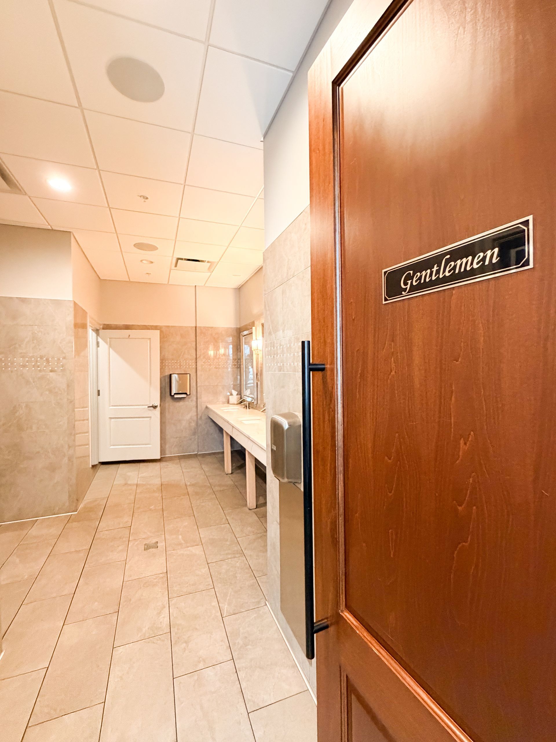 Interior of a men's restroom with a wooden door, sinks, and tiled floor.