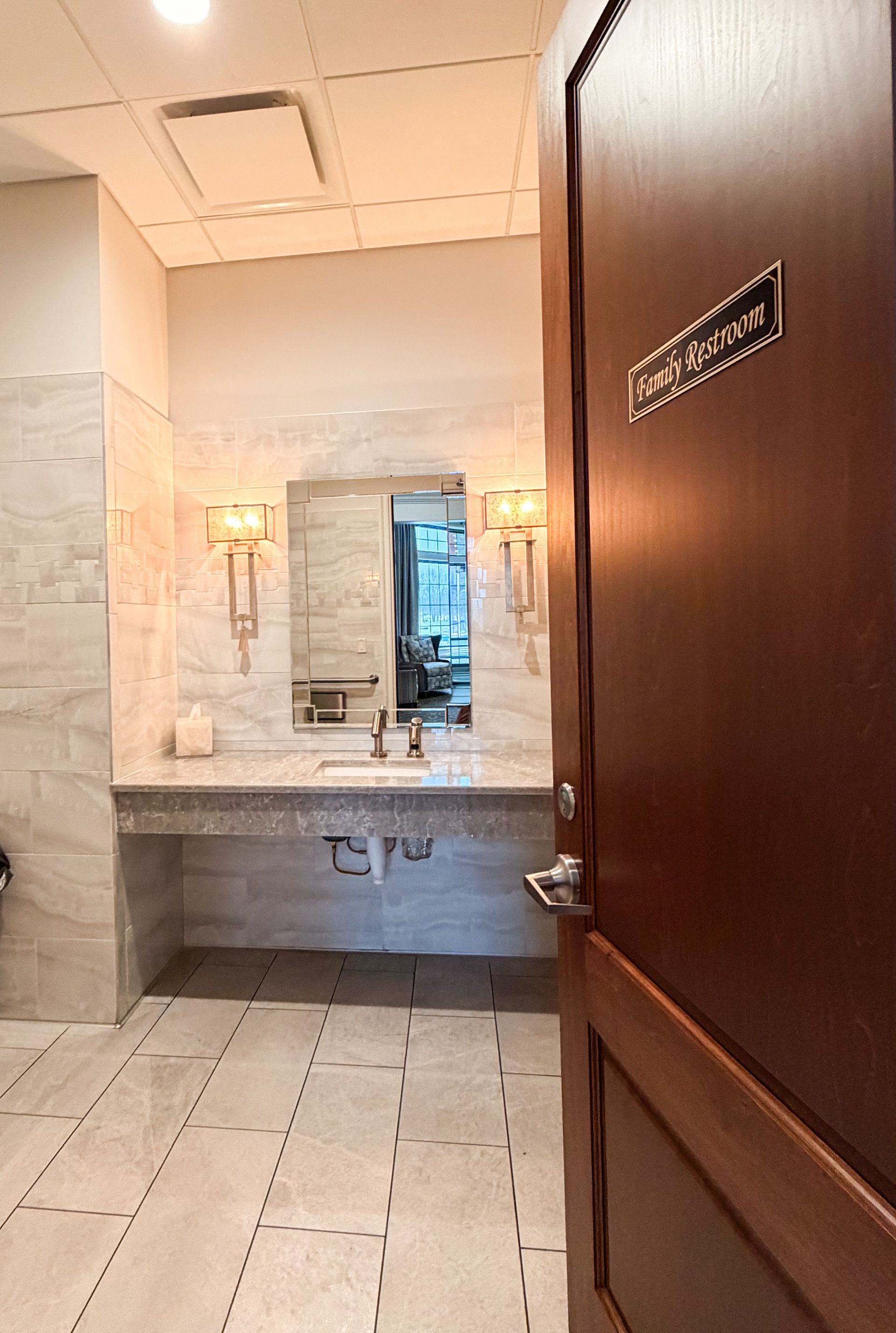 Bathroom with marble walls and countertop, open door, mirror, and sconces. Tile floor.