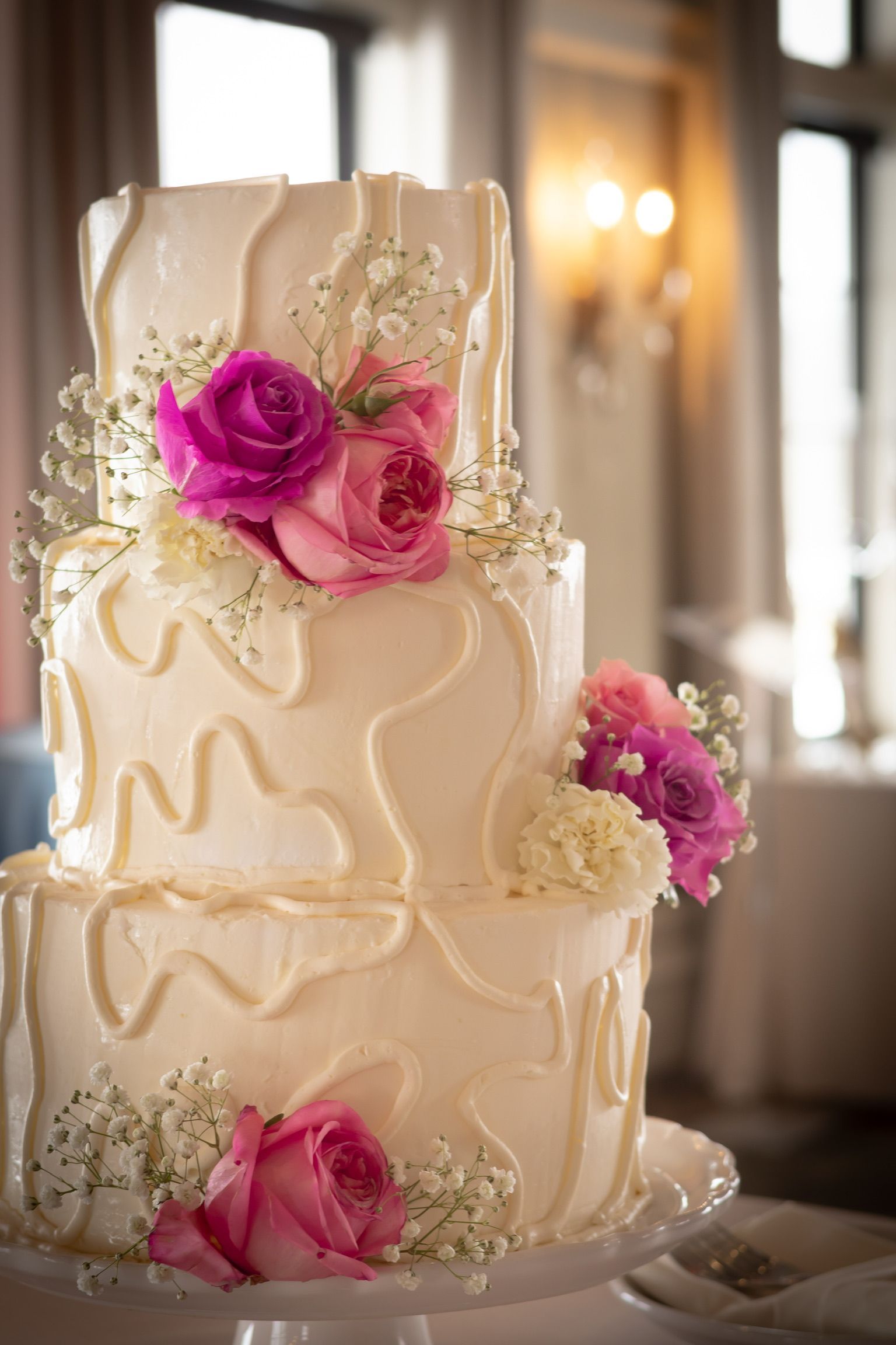 Three-tiered wedding cake with white frosting and pink roses, decorated with white drizzled frosting and baby's breath.