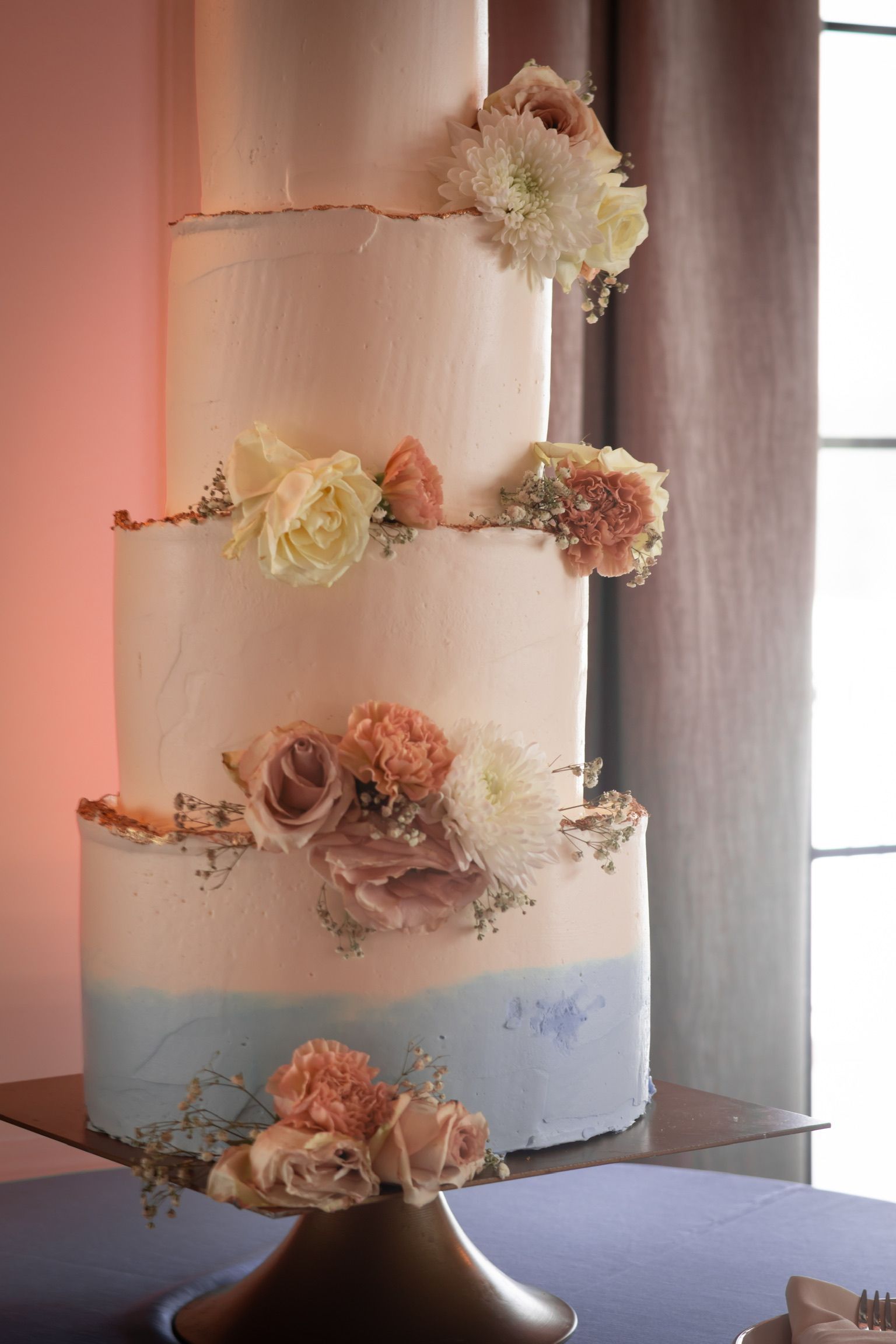 Four-tiered wedding cake with blue ombre, flowers, and gold accents, on a gold cake stand.