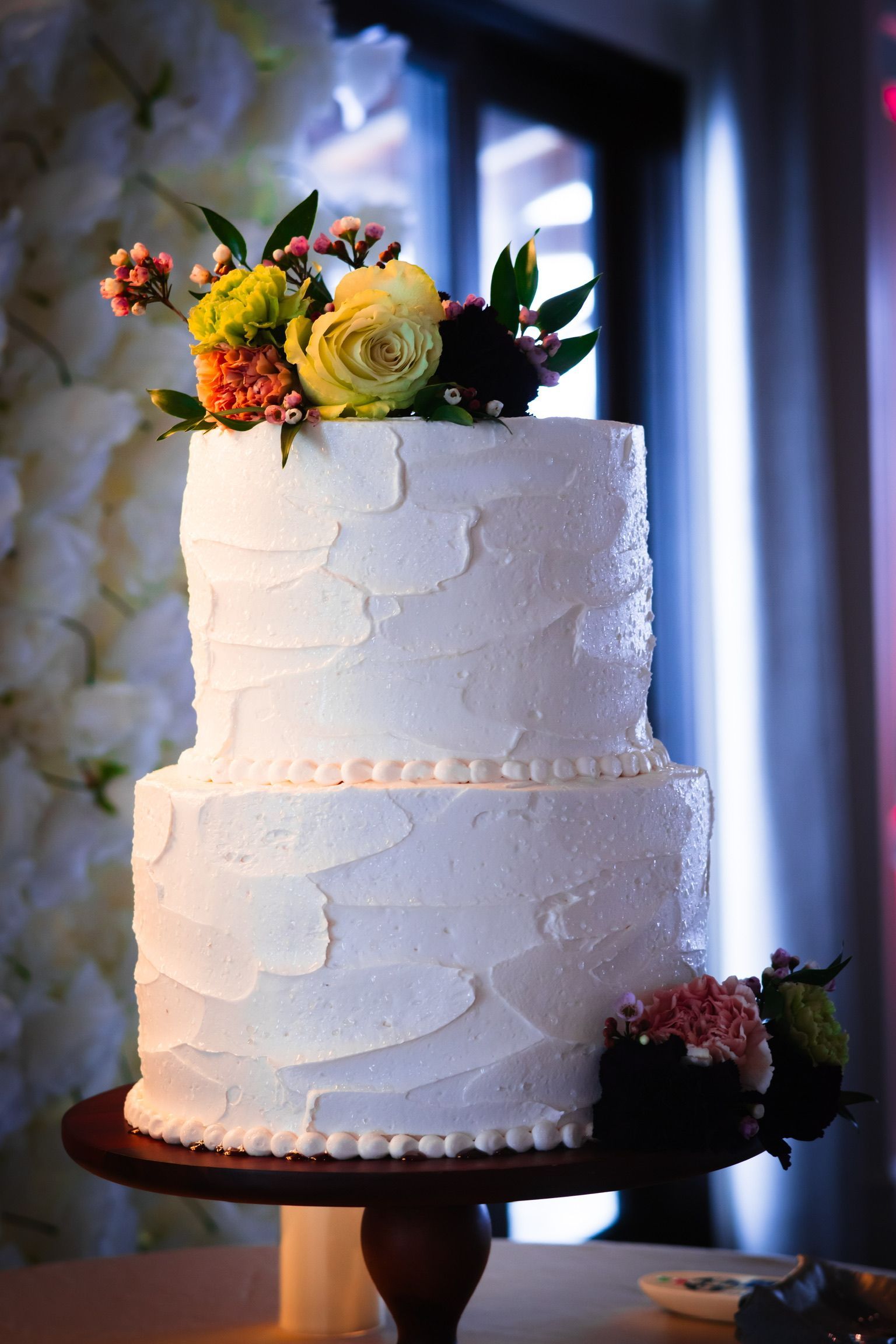 Two-tiered white wedding cake decorated with flowers, on a wooden stand.