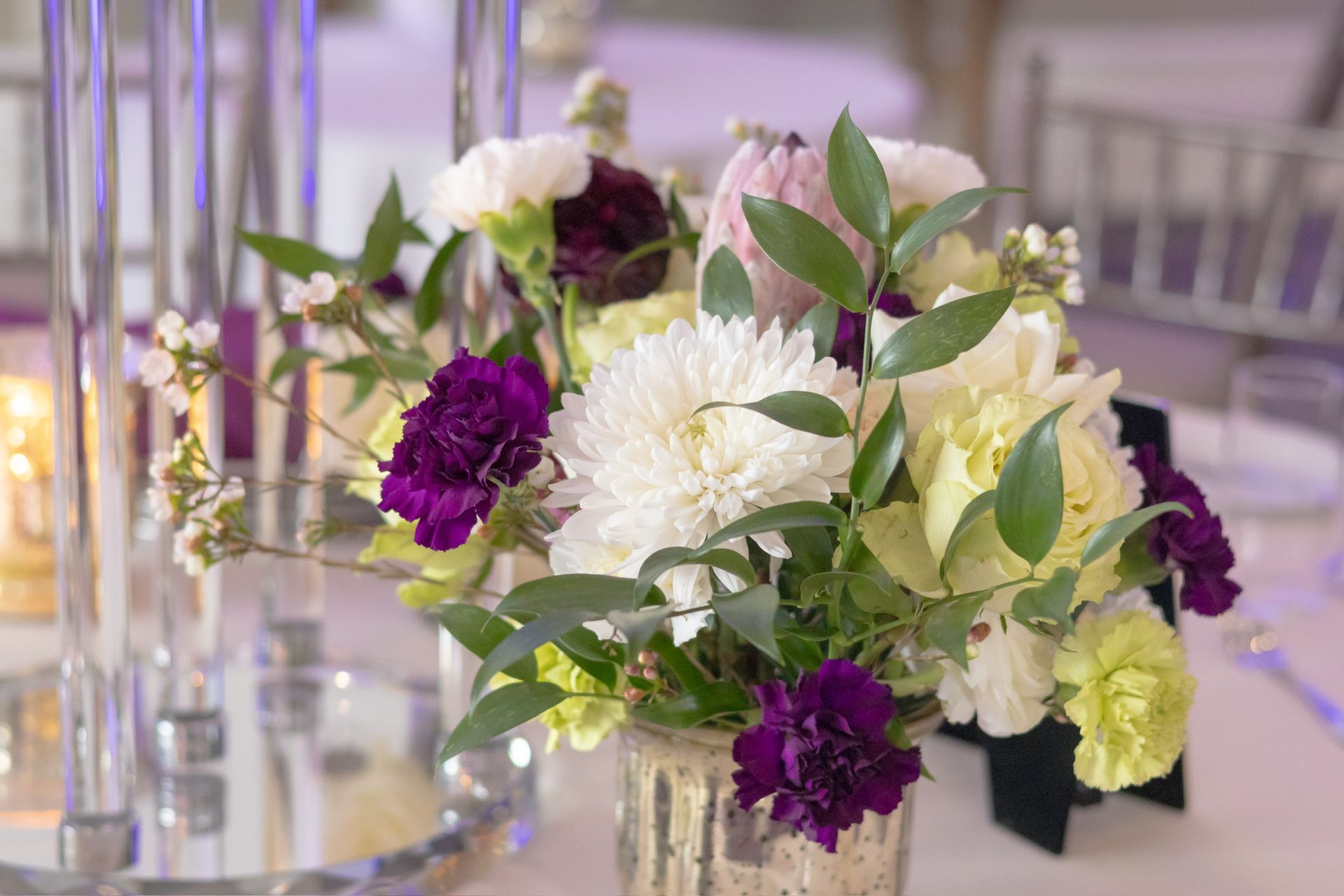 Floral centerpiece with white, purple, and yellow flowers in a gold vase, on a table.