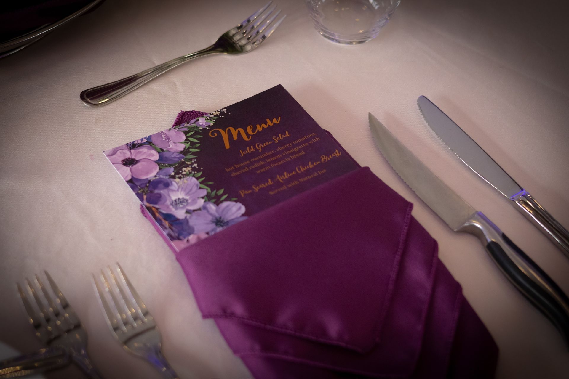 Purple floral menu card in a folded purple napkin on a table with silverware.