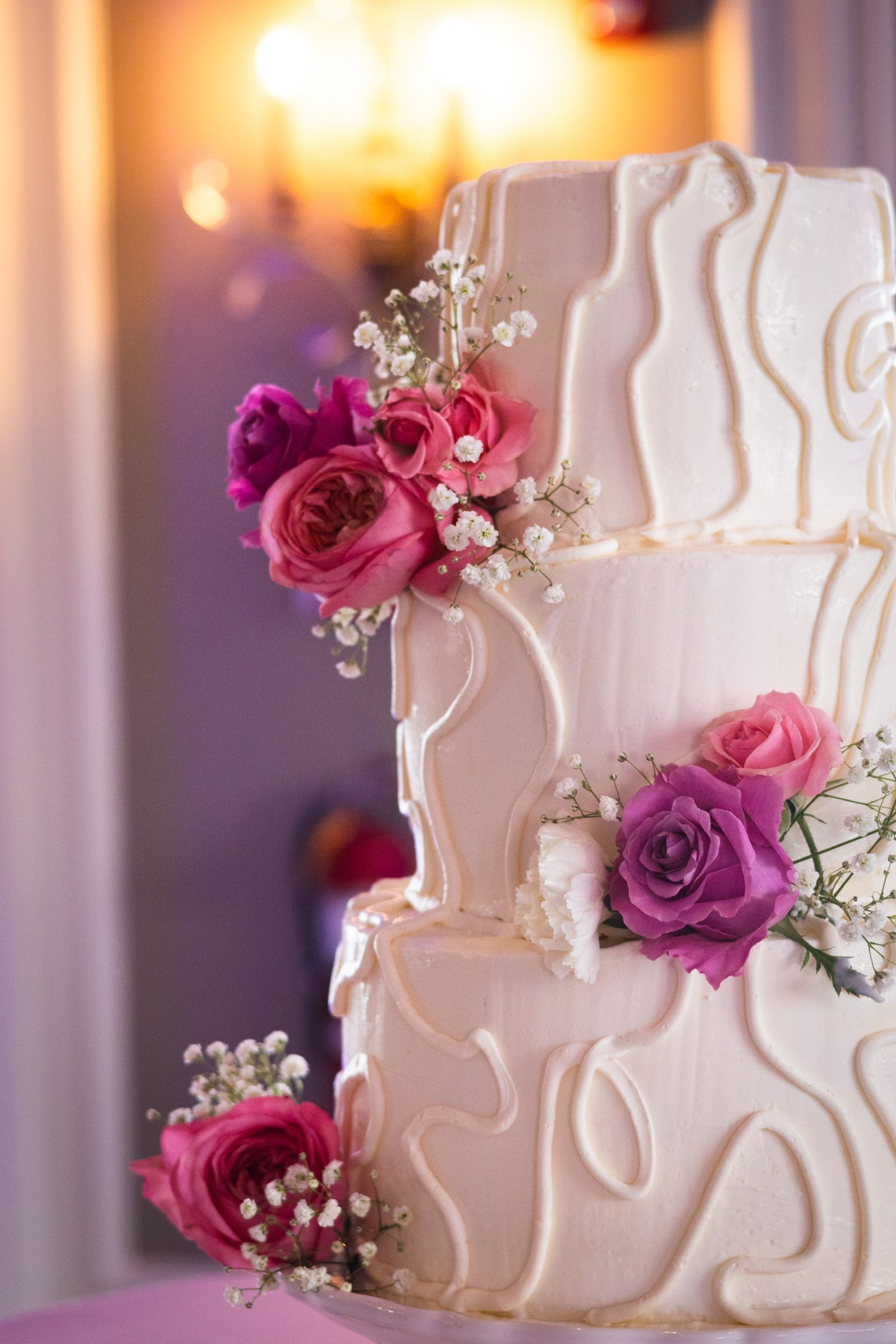 Three-tiered white wedding cake decorated with pink roses and white icing swirls.