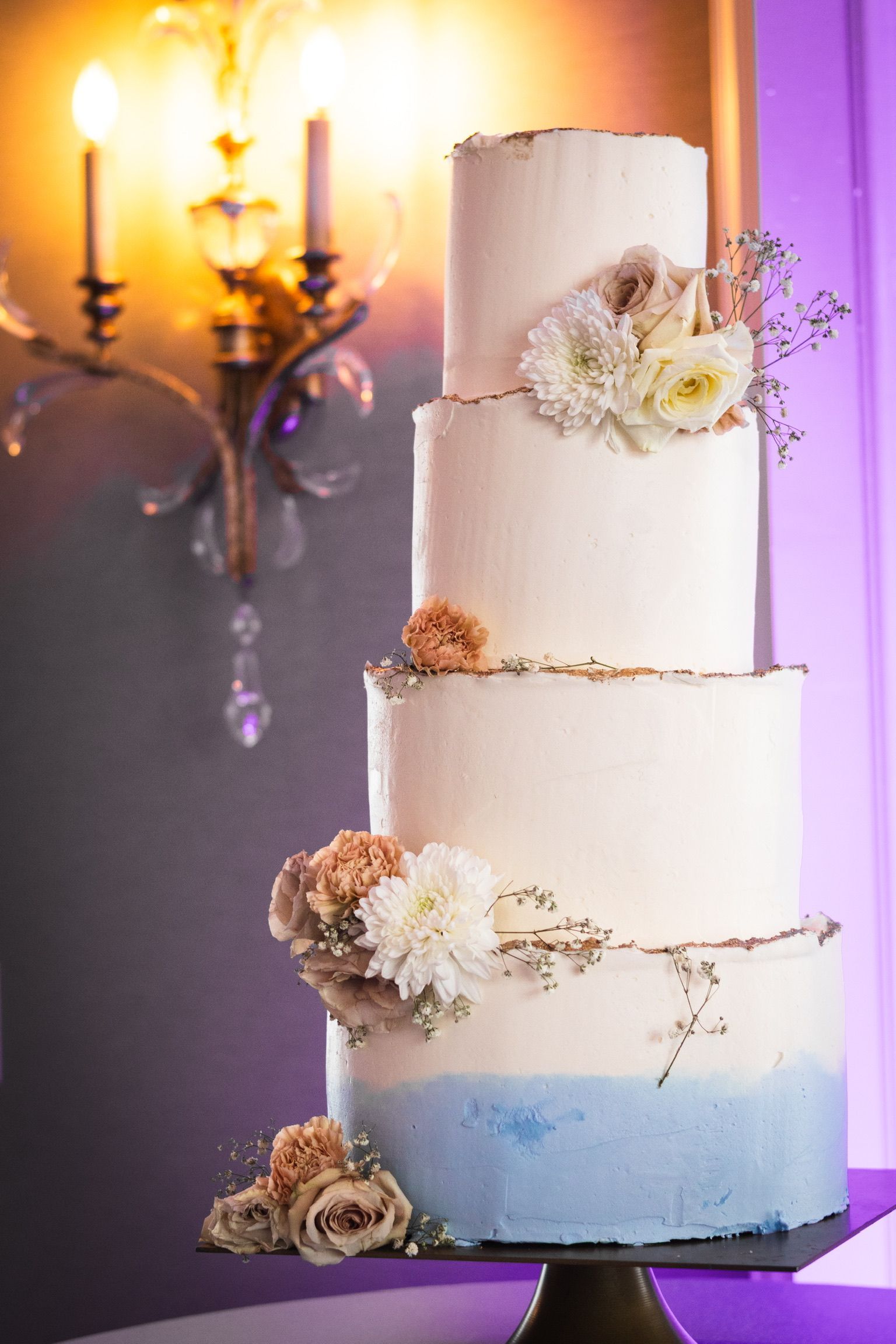 Four-tiered white cake with blue ombre bottom, gold trim, and flower decorations, set against an ornate wall sconce.