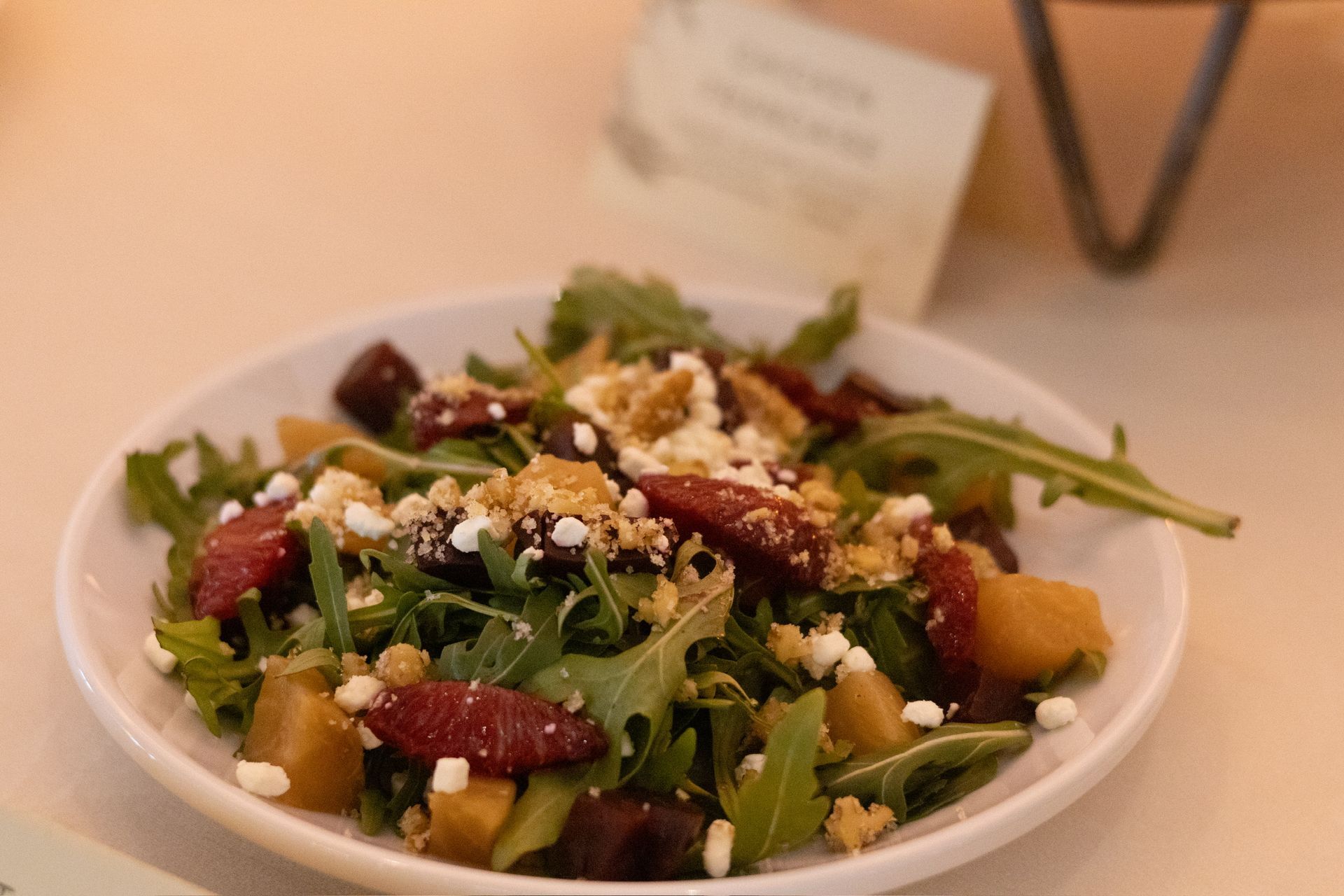 Salad in white bowl with arugula, beets, cheese, and nuts, next to a sign.