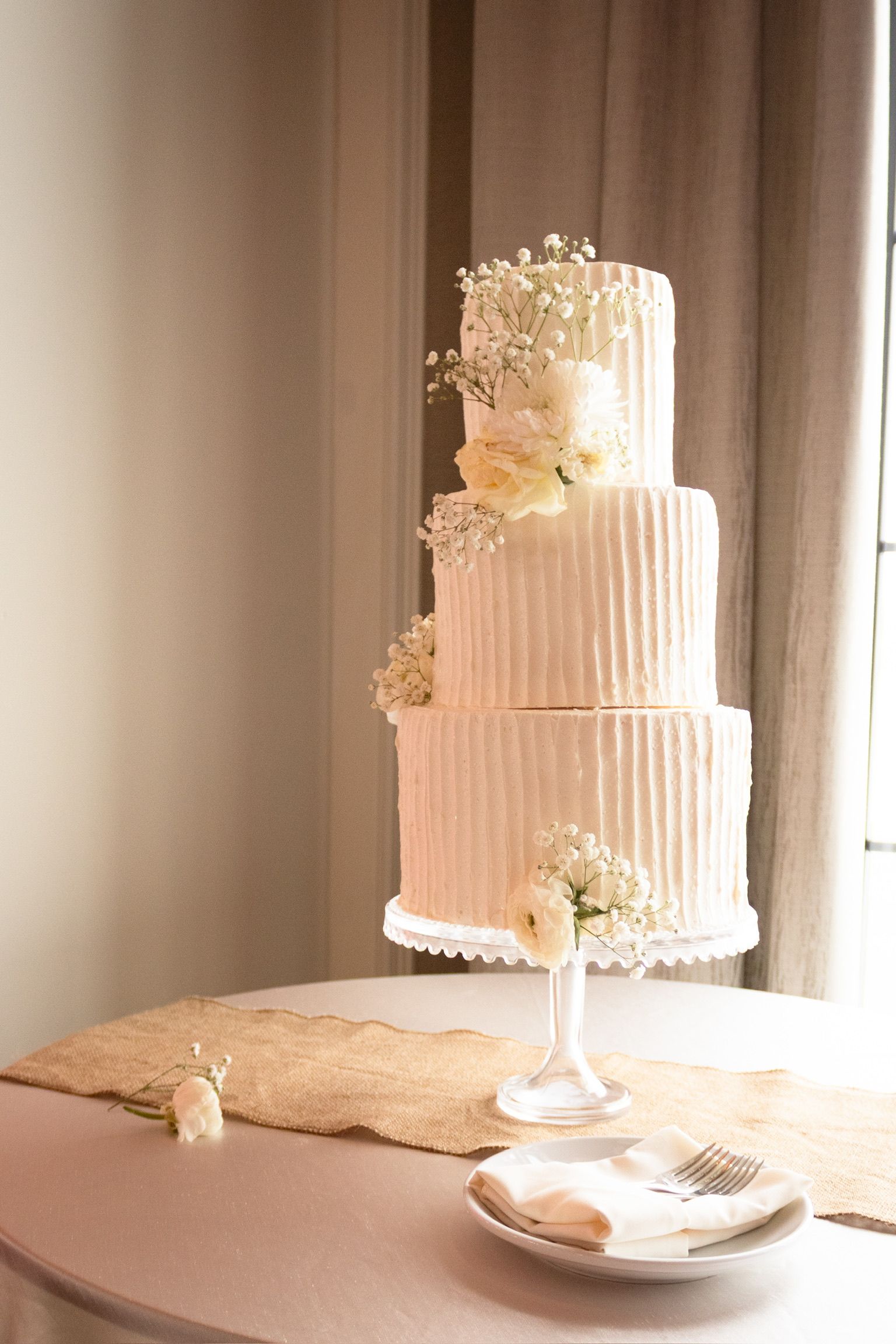 Three-tiered wedding cake, off-white with vertical texture, decorated with white flowers, on a table.