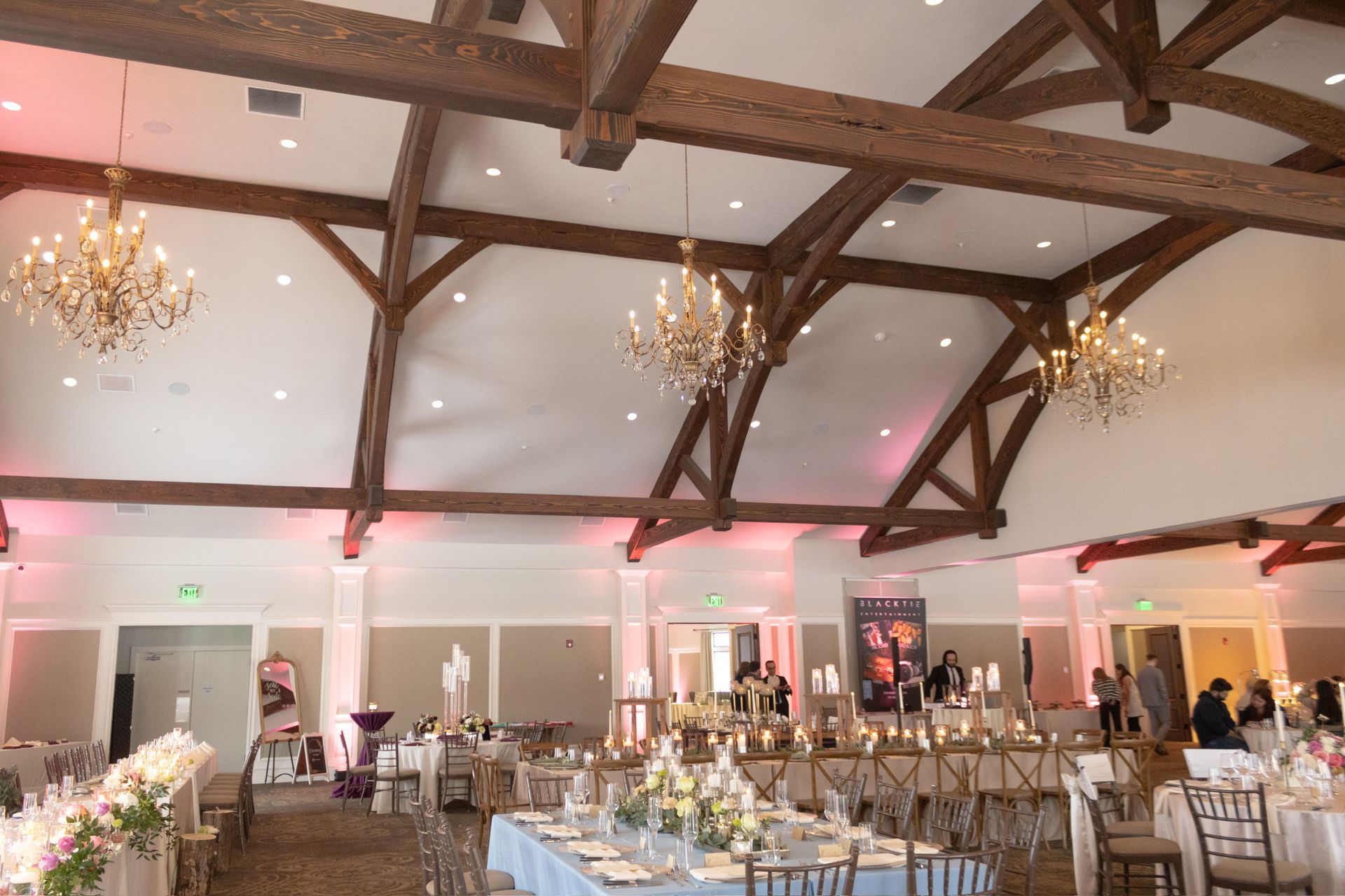 Banquet hall with tables set for a gathering, chandeliers, wooden beams, and uplighting.