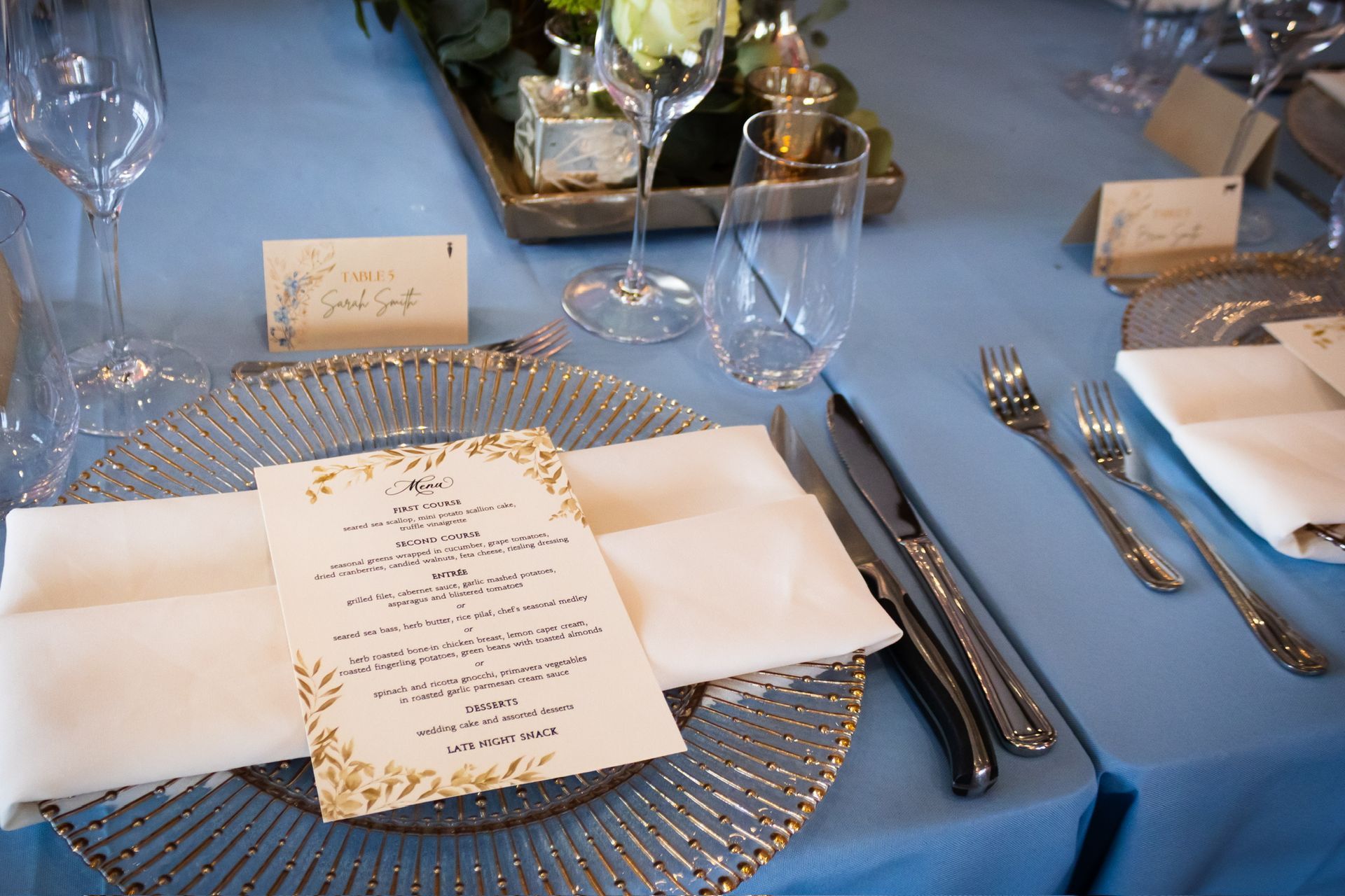 Table setting with blue tablecloth, gold-rimmed glasses, menu card, and silverware.