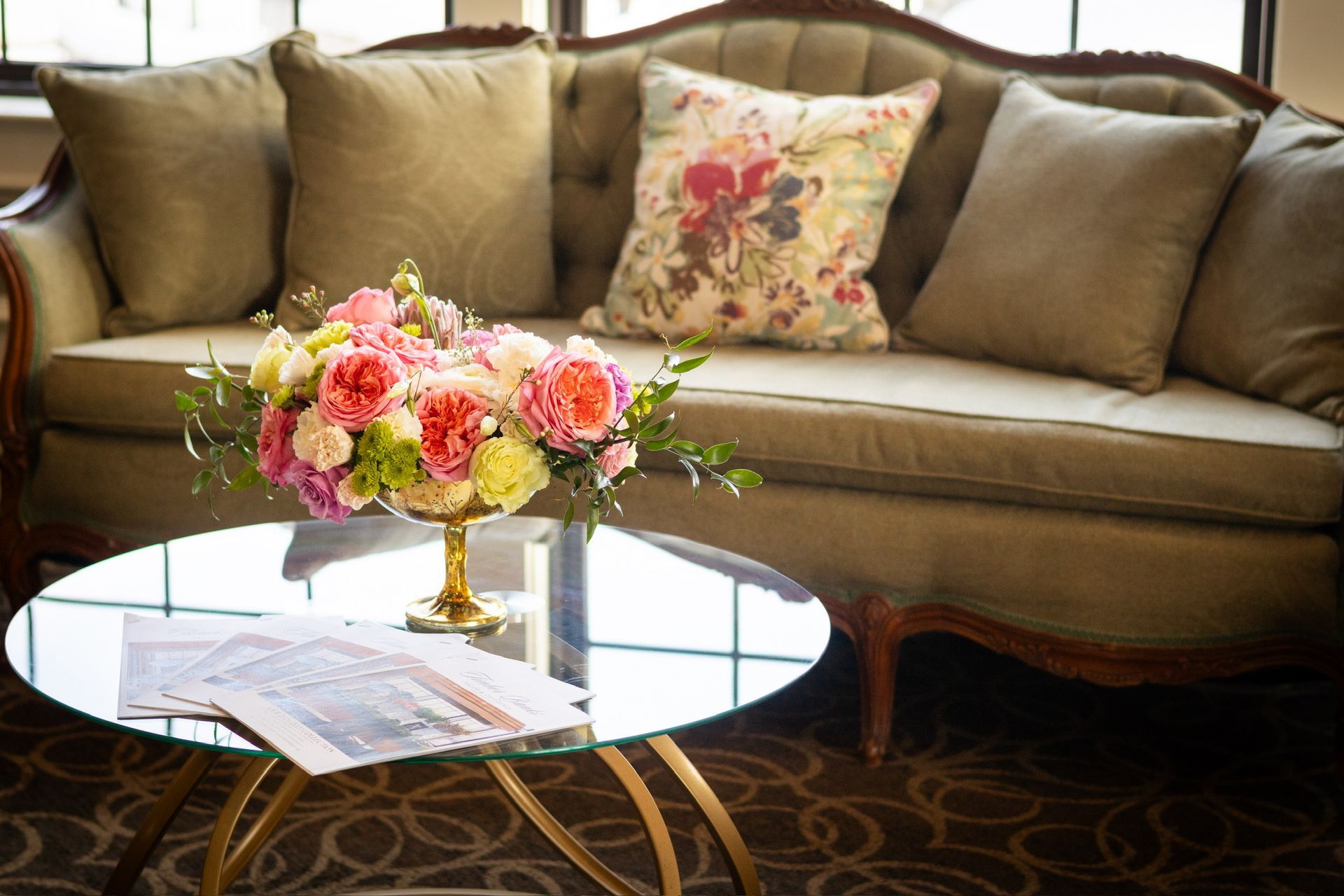 Round glass coffee table with floral arrangement and magazine, in front of a green couch with pillows.