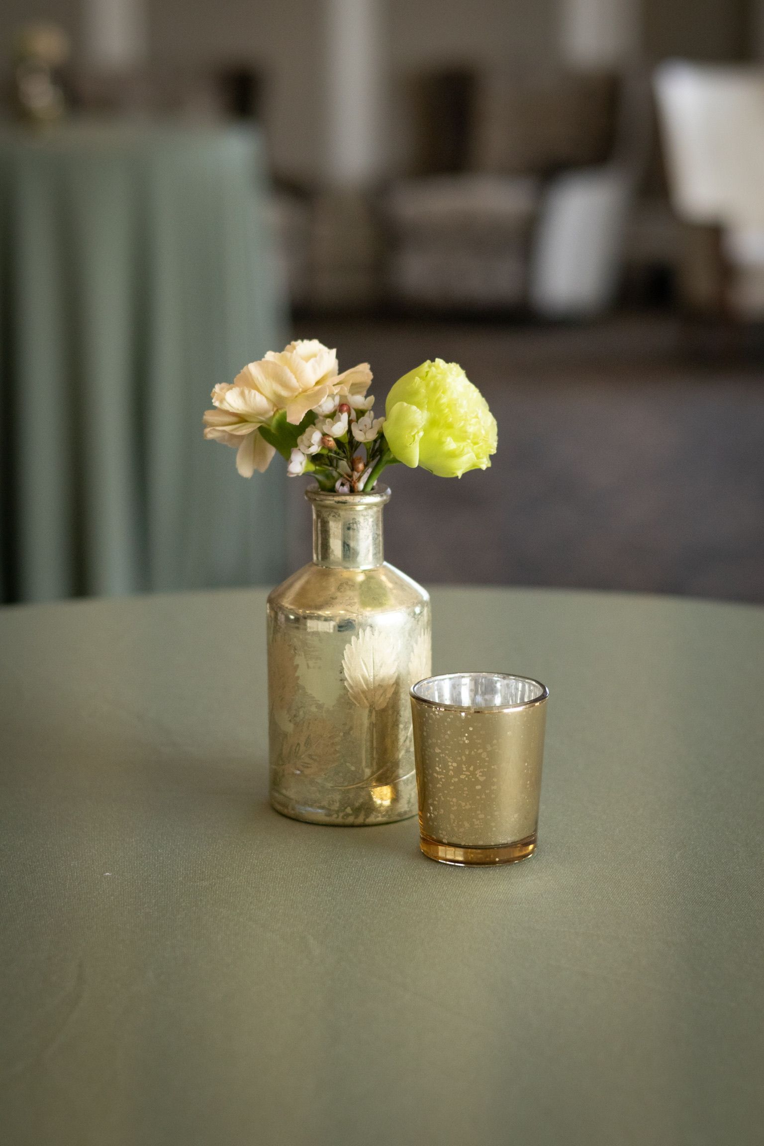 Small gold vase with flowers and a candle on a round table covered in green fabric.