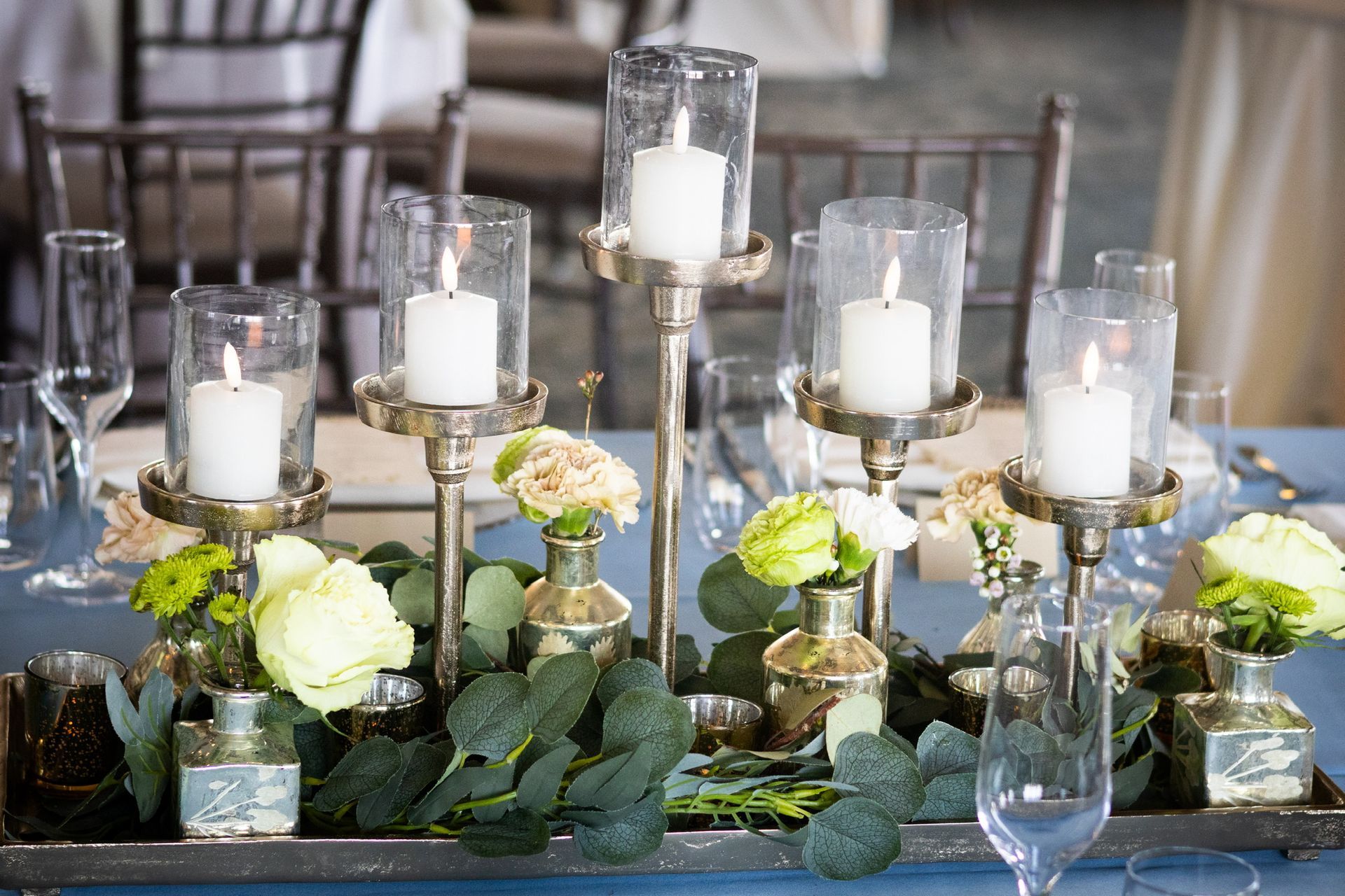 Table centerpiece with candles, flowers, and greenery on a blue tablecloth; elegant dining setting.