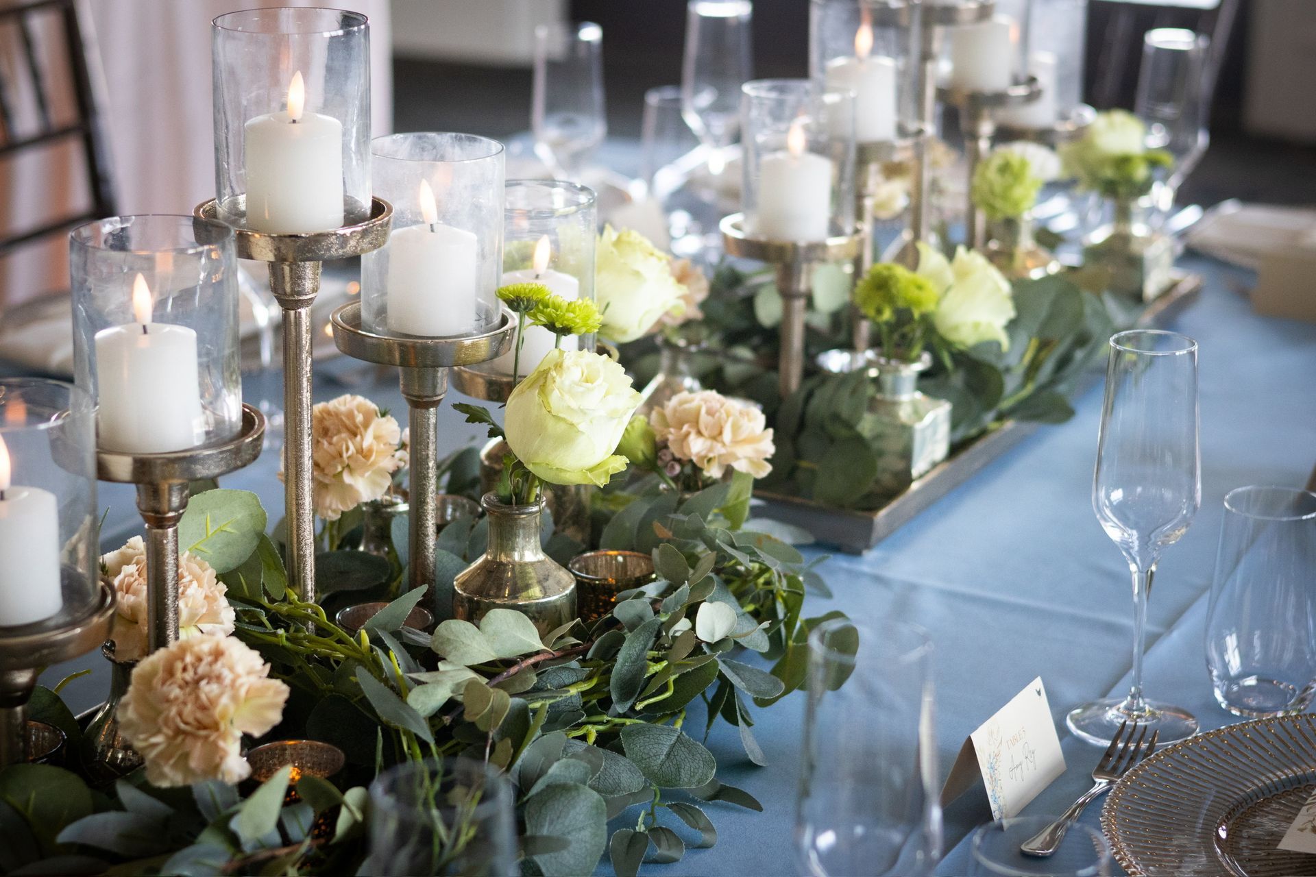 Elegant table setting with candles, flowers, and glasses on a light blue tablecloth.