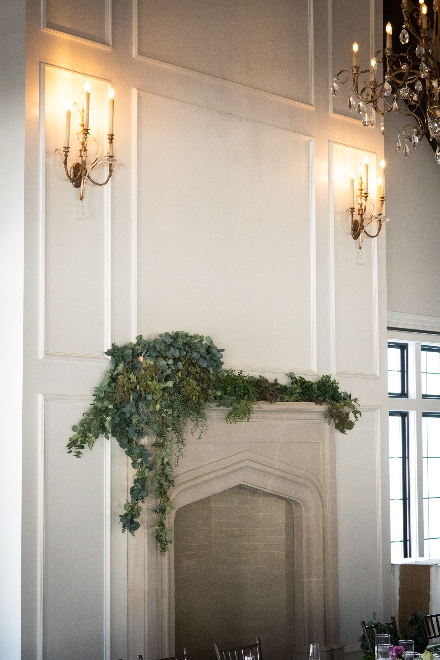 Ornate fireplace with greenery and sconces on a white wall.