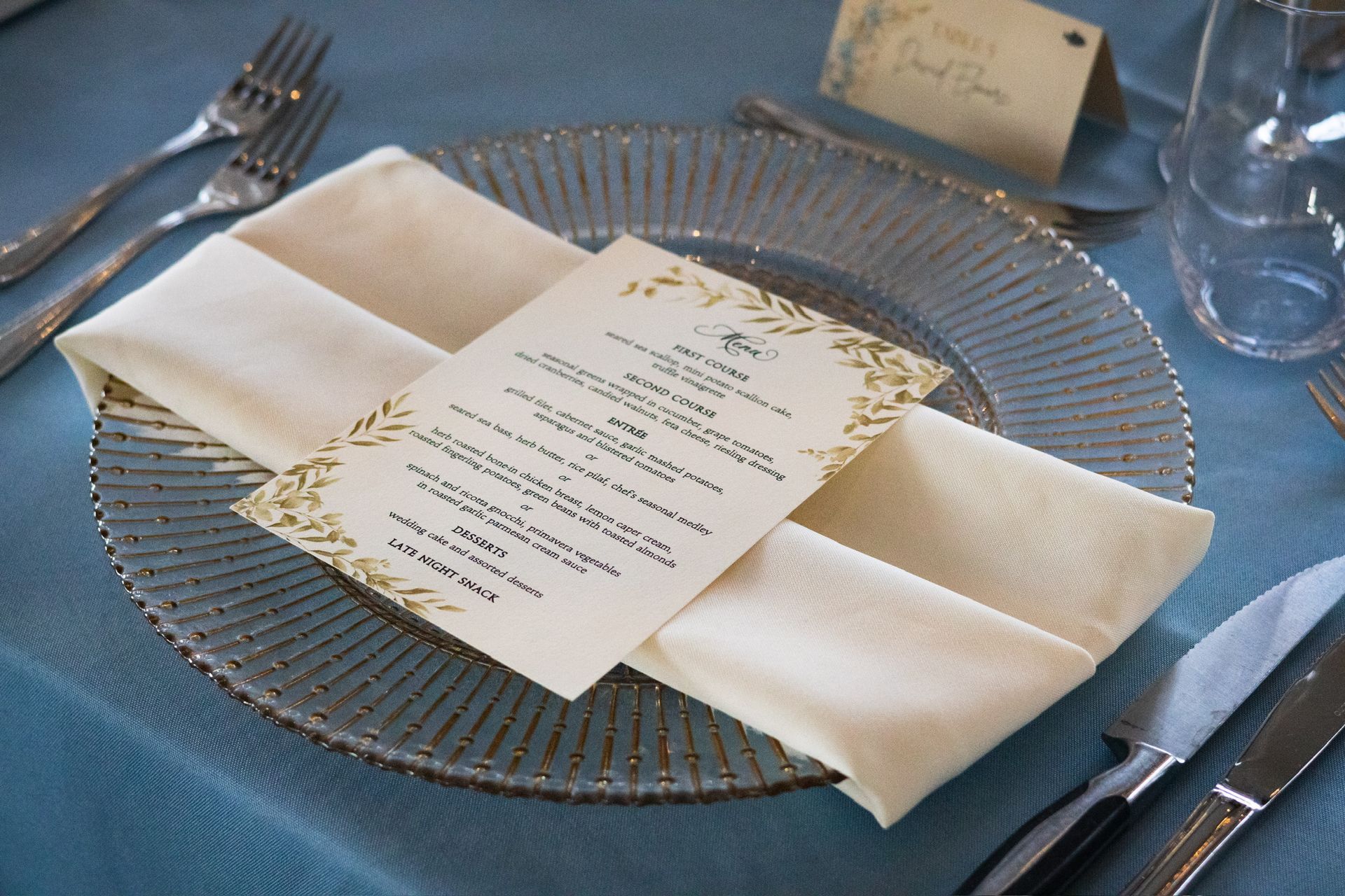 Place setting with a menu card, folded napkin, and silverware on a blue tablecloth.
