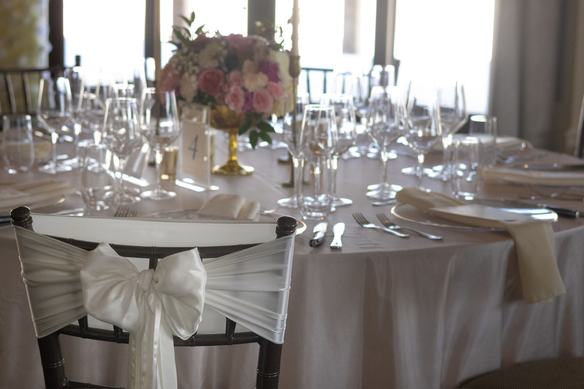 Formal dining table set with white tablecloth, glassware, and floral centerpiece; chair with white bow.