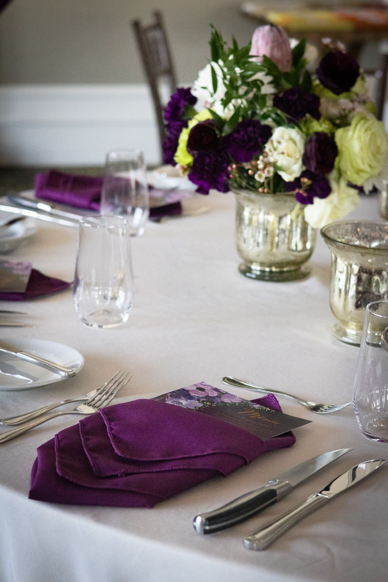 Formal dining table set with purple napkins, silverware, and floral centerpiece.