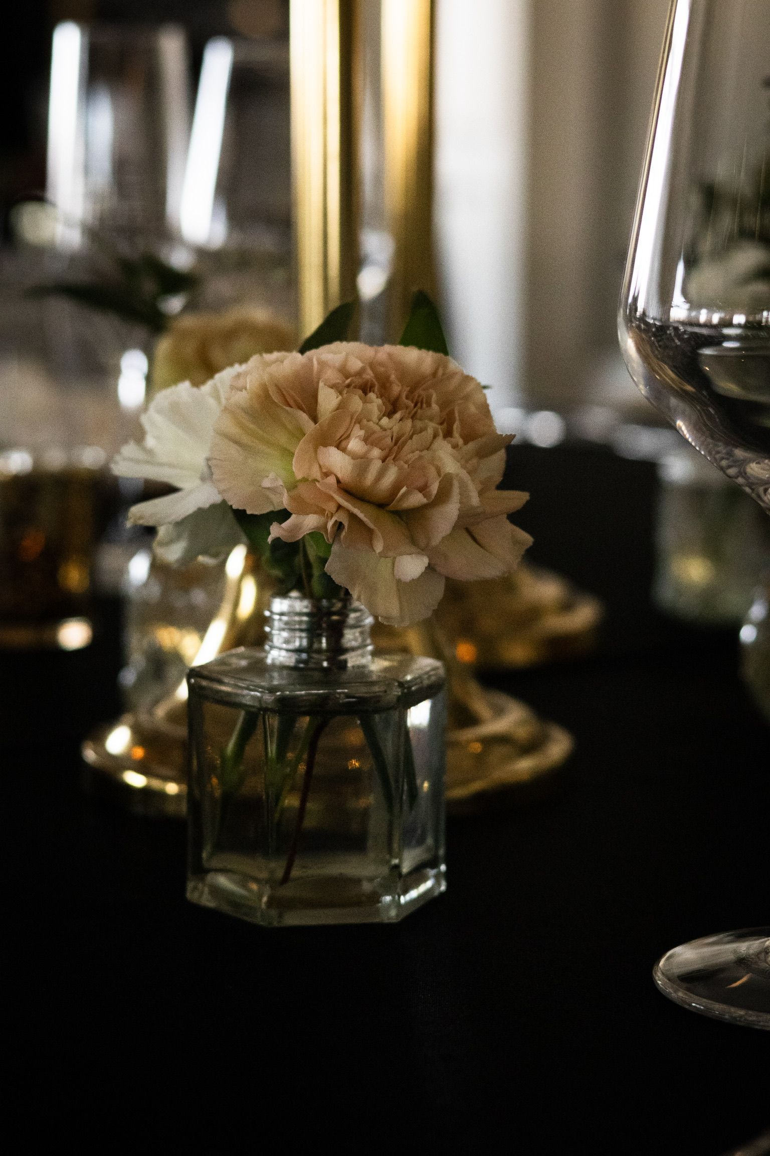 A small beige and white flower in a glass vase on a table, with gold candle holders in the background.