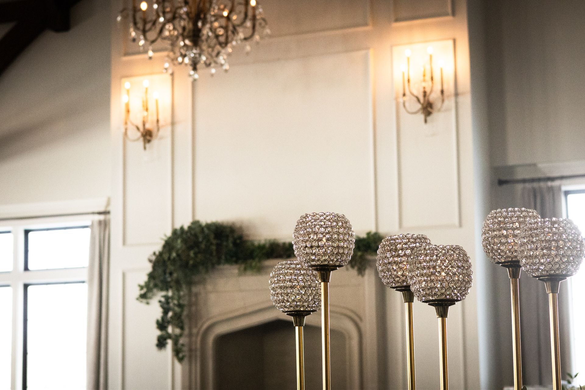 Crystal globes on gold stands in front of a fireplace with green garland, chandelier, and sconces.
