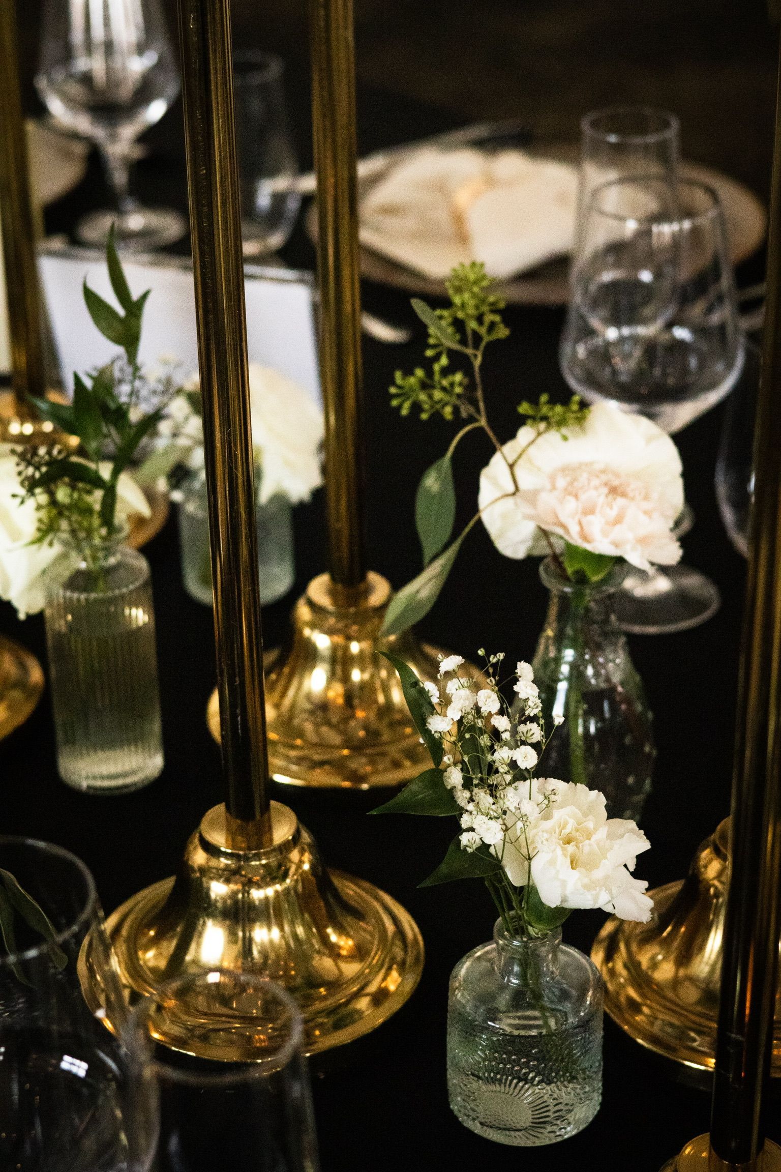 Golden candle holders and small floral arrangements on a black table with glassware.