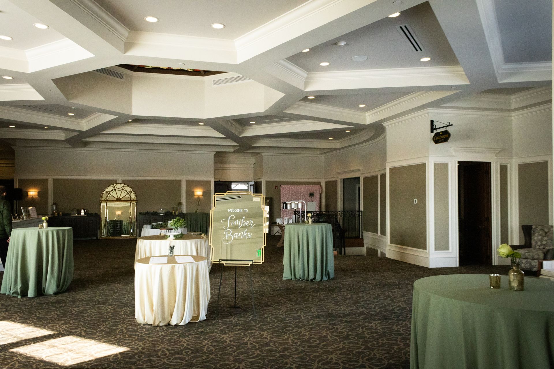 A light-filled event room with round tables covered in green and cream tablecloths. Sign and mirrors are present.