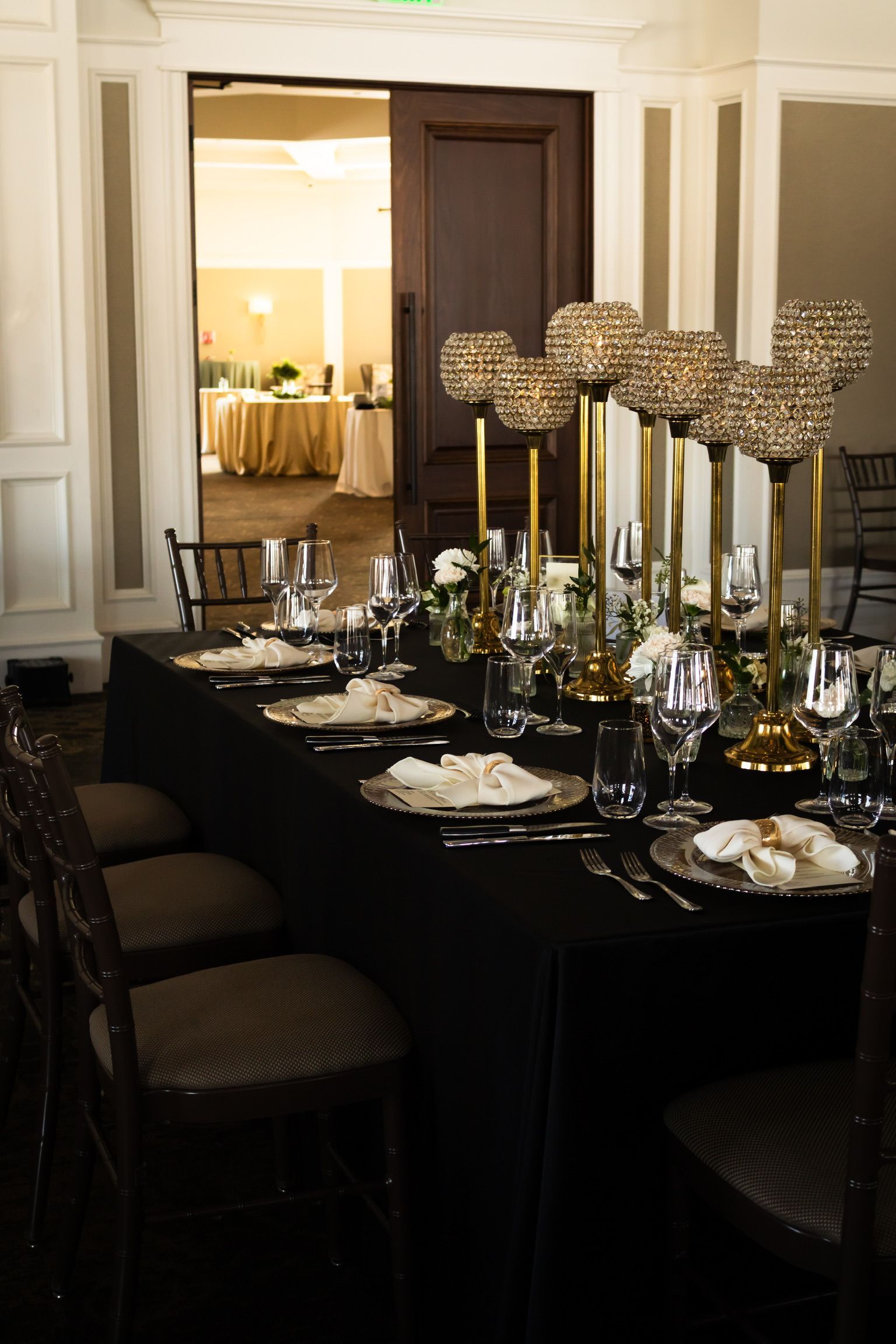 Formal dining table set for a special event, with black tablecloth, gold accents, and tall centerpieces.