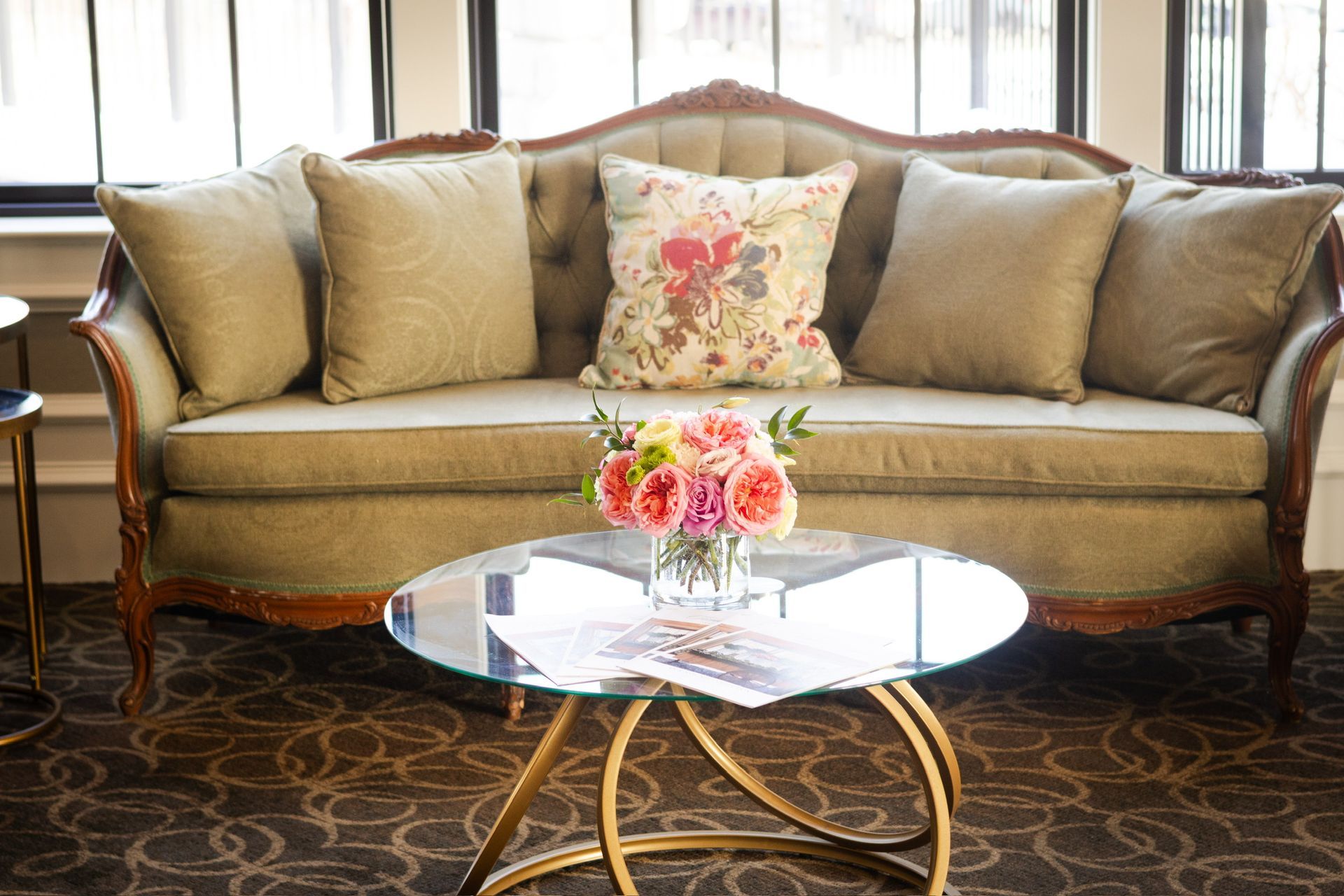 A tufted olive green couch with decorative carved wood, floral pillow, glass coffee table, and bouquet.