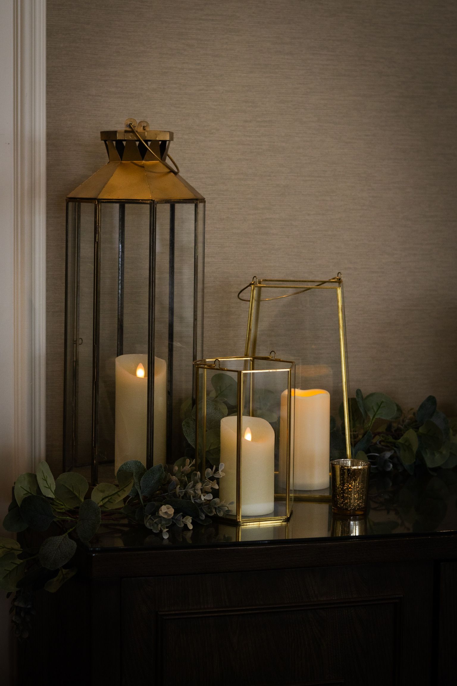 Lanterns with lit candles on a dark surface, greenery, against a textured wall.