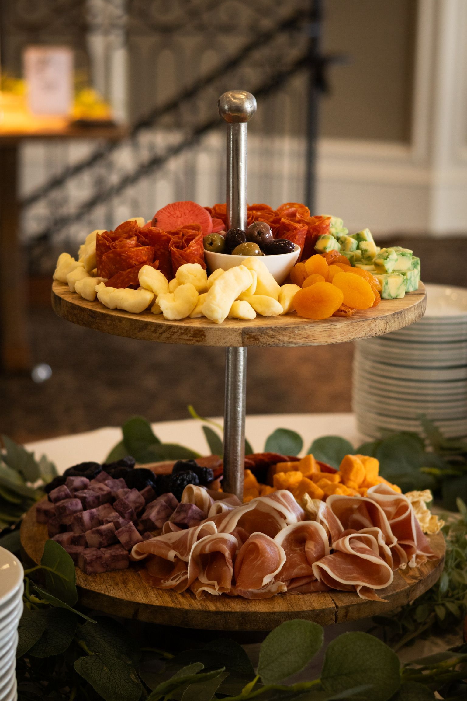 Two-tiered wooden platter with charcuterie, cheese, olives, and fruit.