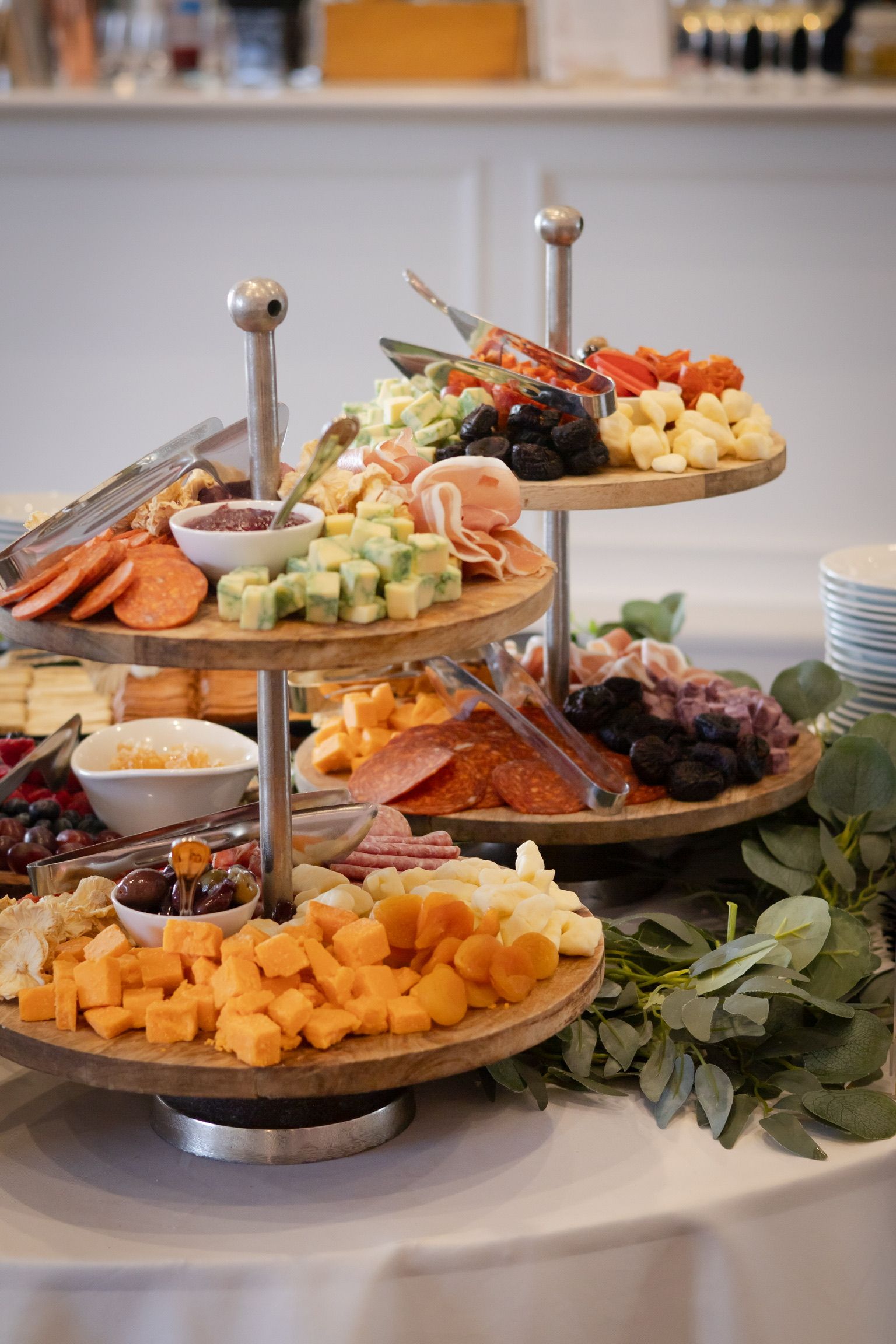 Three-tiered food server holding cheese, meats, crackers, and olives on a white-covered table, greenery in the corner.