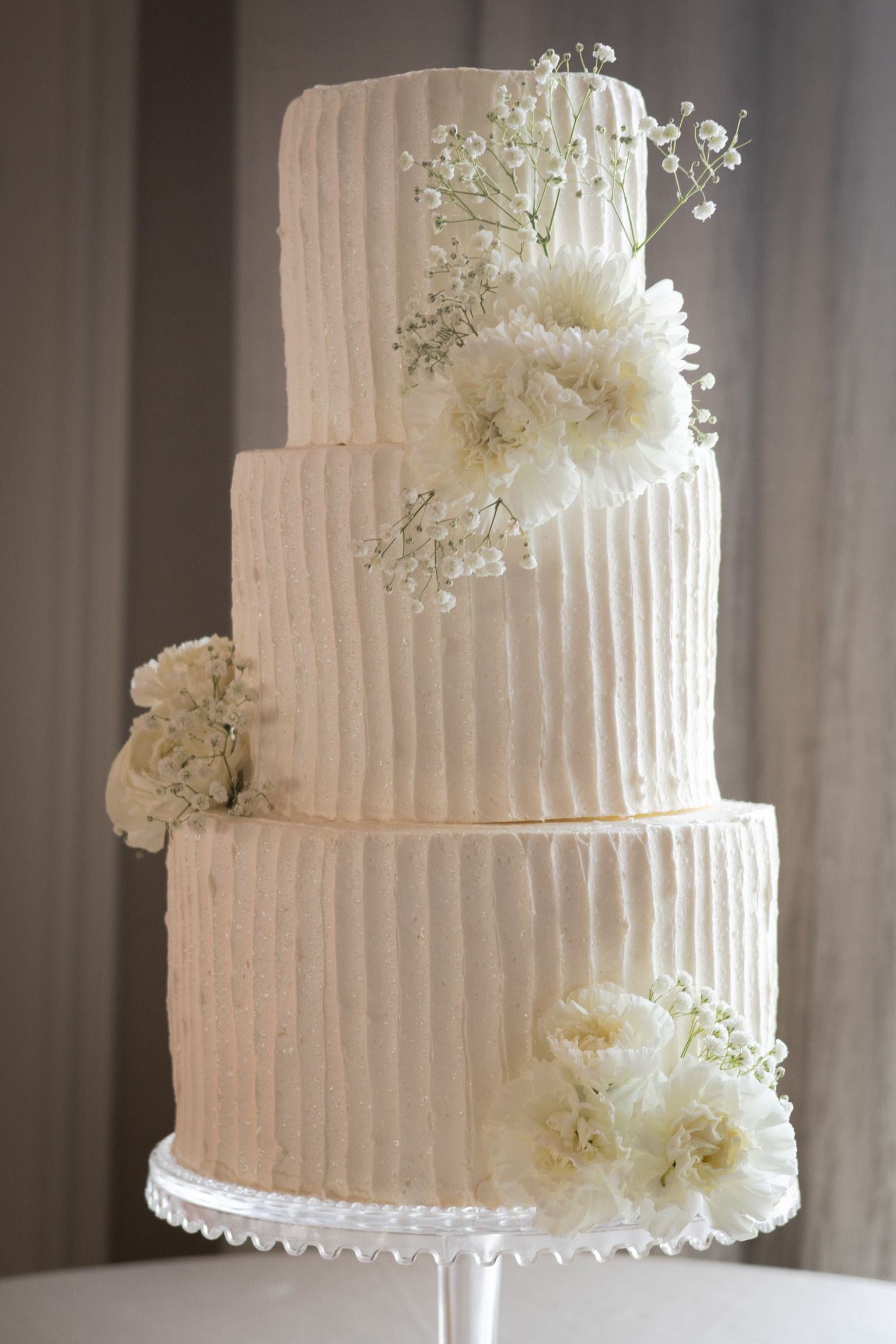 Three-tiered white wedding cake with textured frosting and white floral accents on a clear cake stand.