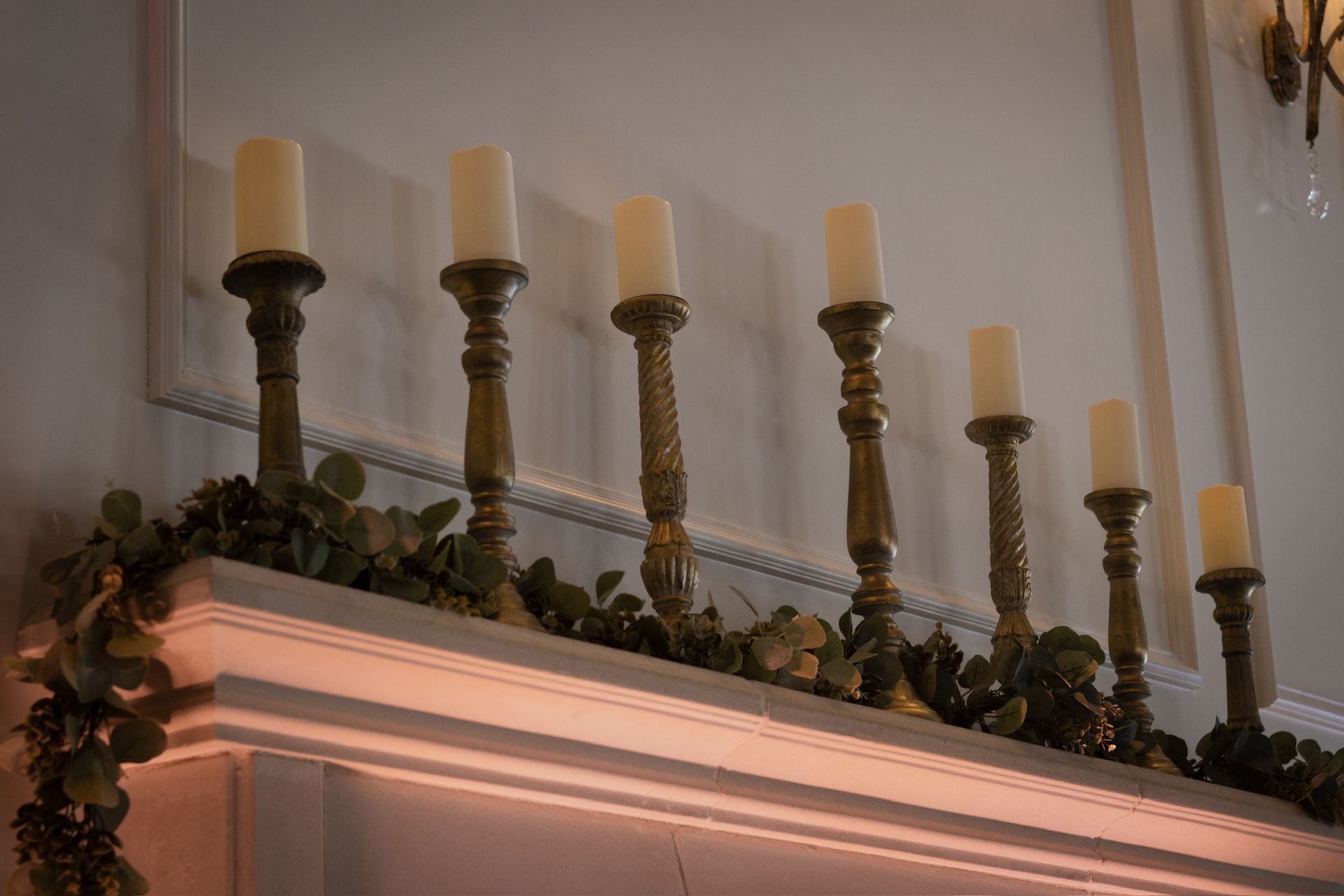 Candle holders with white candles atop a white mantle decorated with greenery.