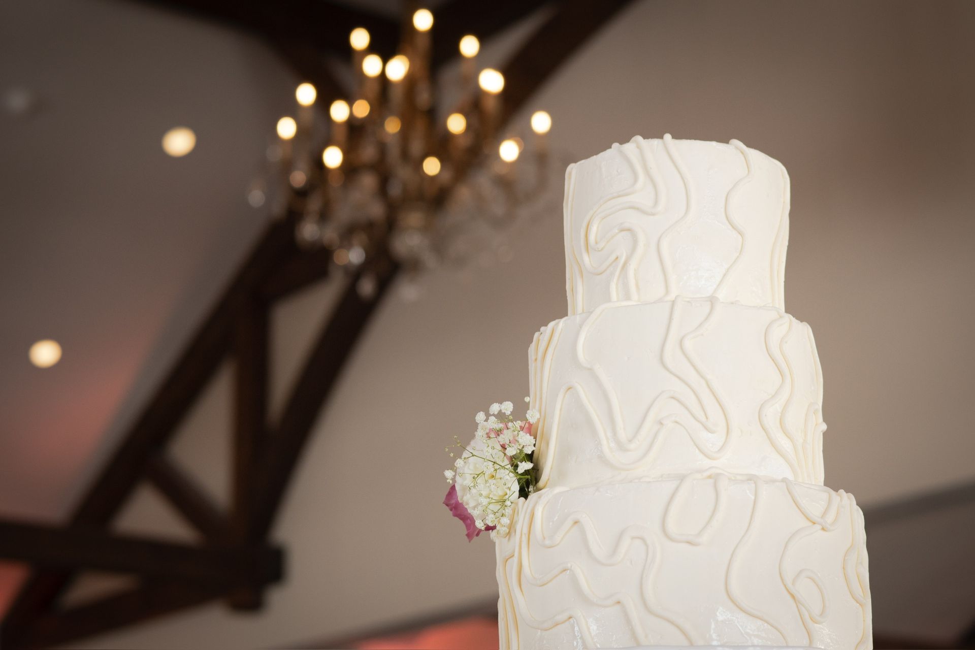 Three-tiered white wedding cake with textured frosting and flowers, with a chandelier and exposed beams in the background.