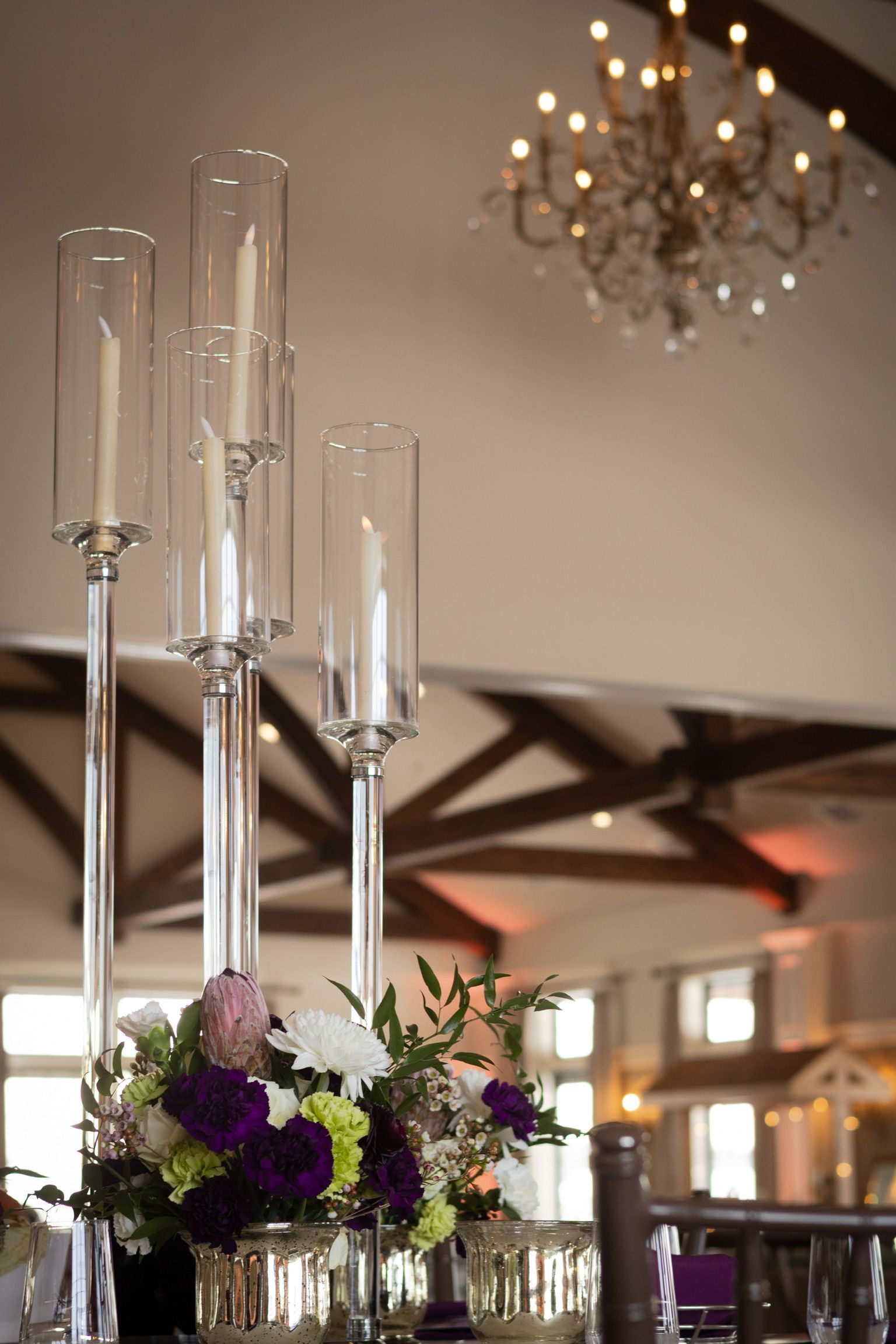 Centerpiece with tall glass candle holders and floral arrangement. Chandelier and wood beam ceiling in the background.