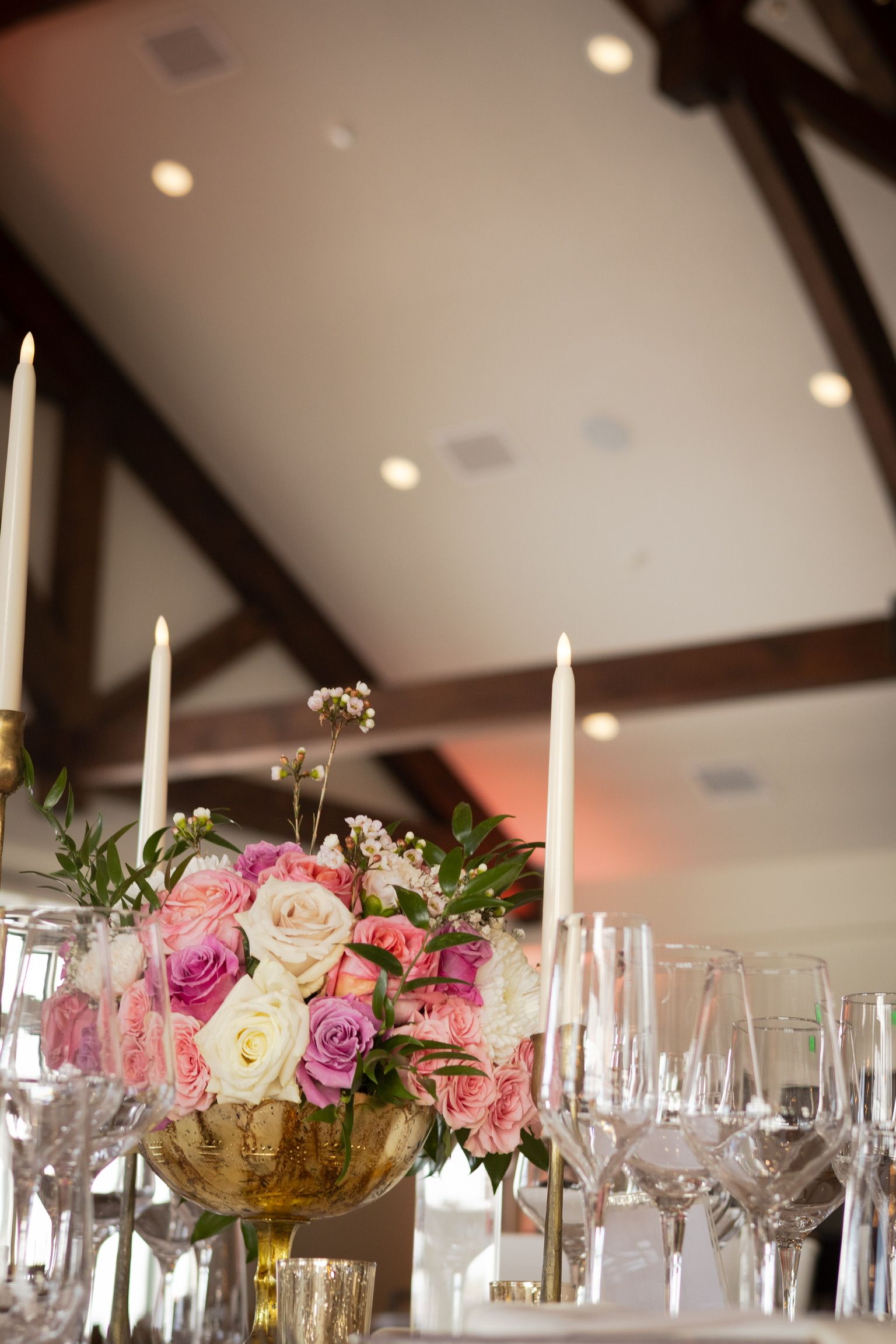 Floral centerpiece with pink and white roses in a gold bowl, candles, and glasses set for a formal event.