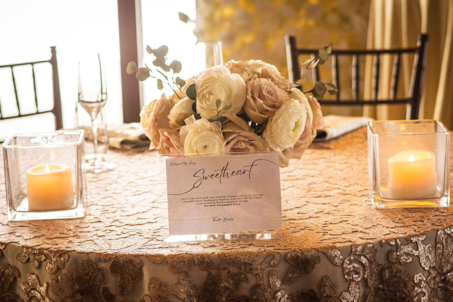 Centerpiece on a table: floral arrangement with a sign, flanked by candles in glass holders.