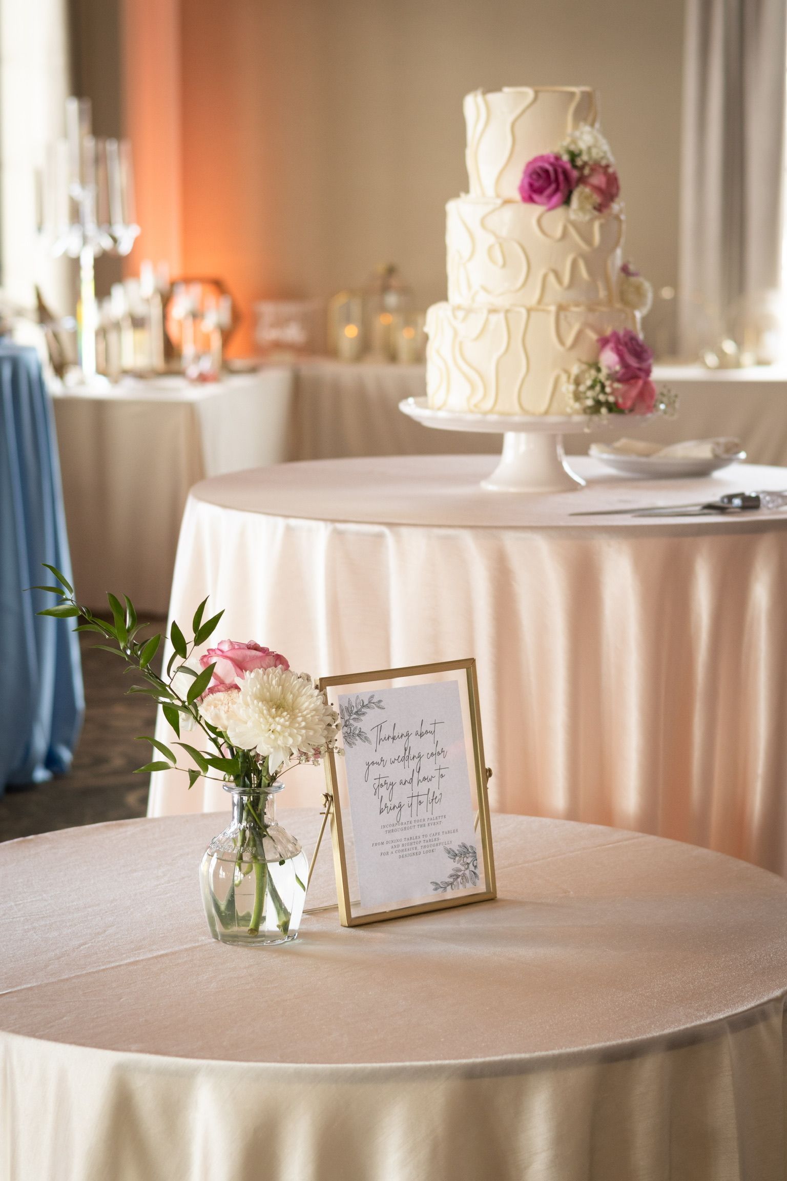 Wedding cake on a round table, with a floral arrangement and framed sign. Pink and white tones.