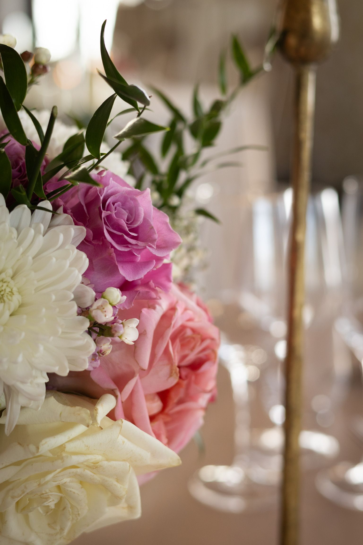 Close-up of a floral arrangement with white and pink roses on a table, partially obscuring wine glasses.