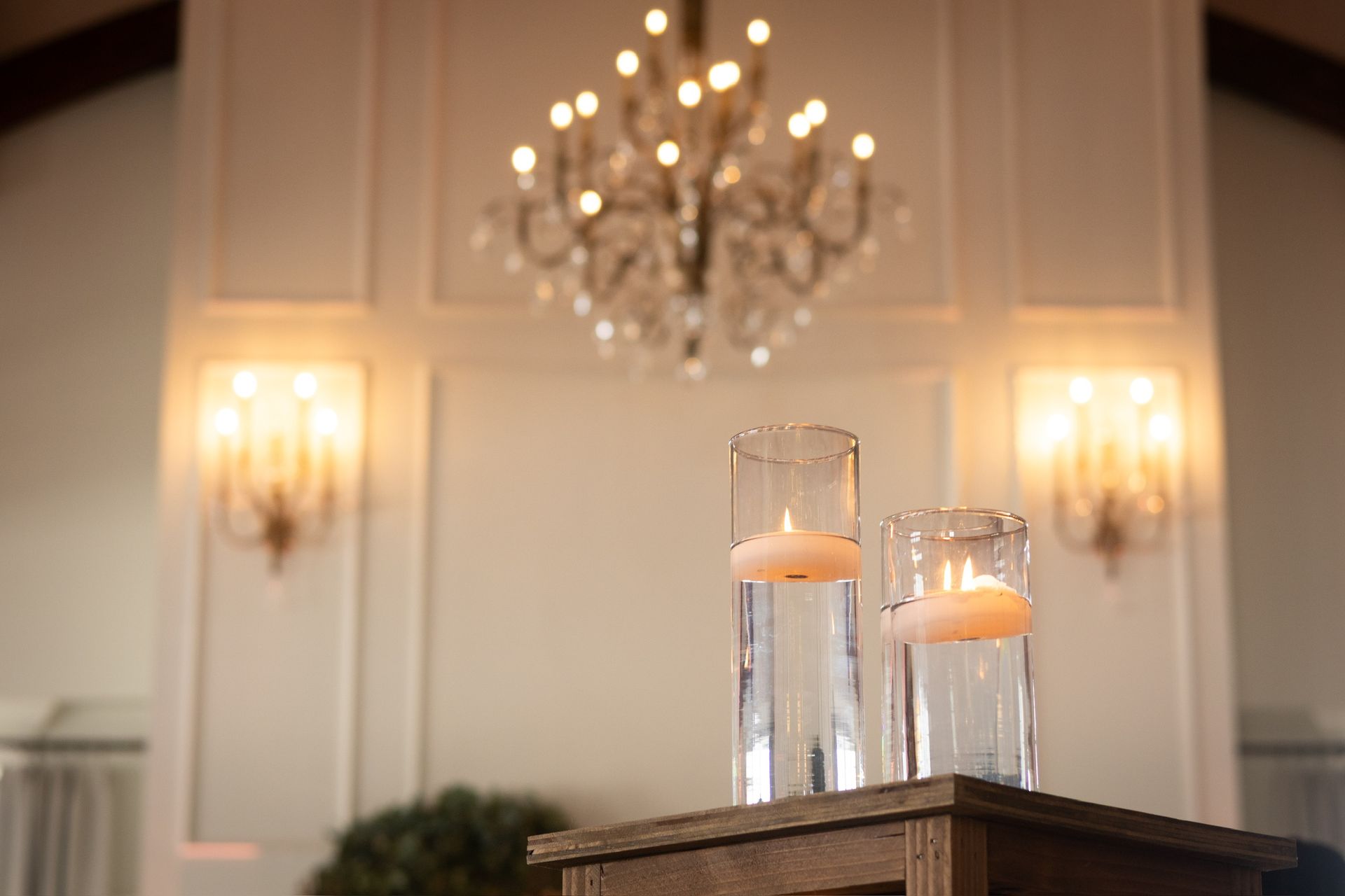 Two glass cylinders with floating candles on a wooden stand, in front of a white wall with chandelier and sconces.