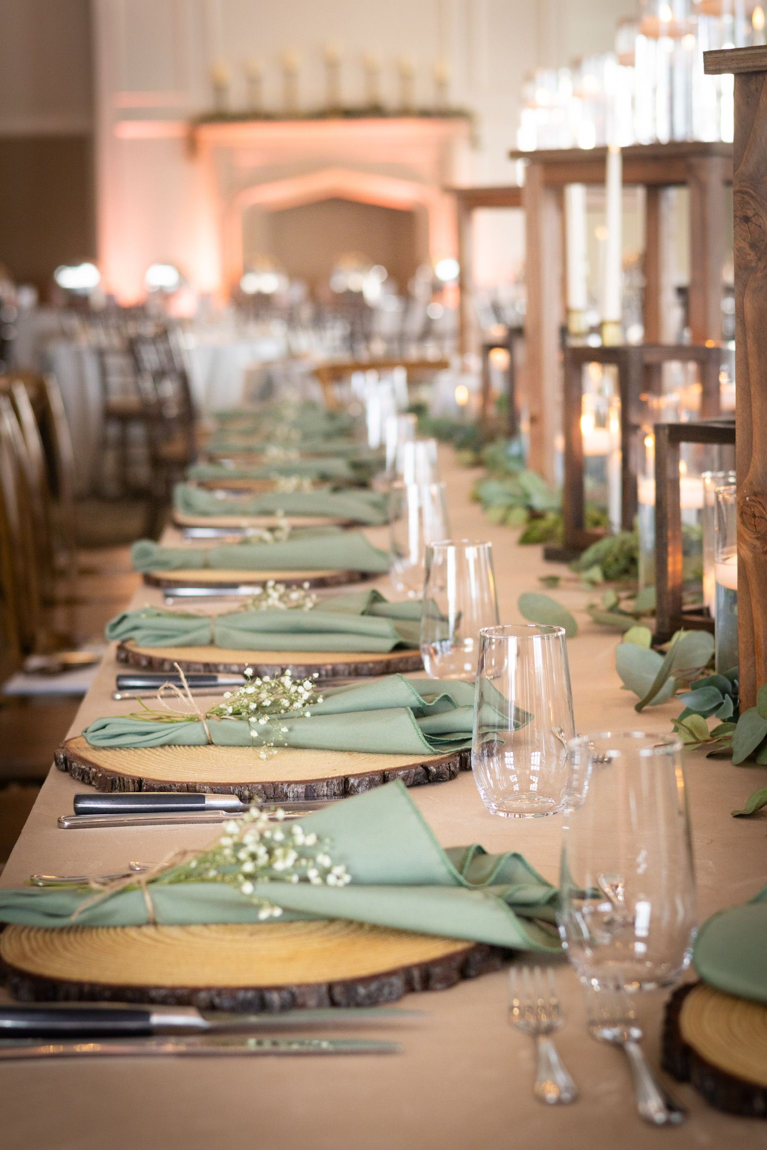 Long wooden table set for a wedding reception with green napkins, wood chargers, and greenery.