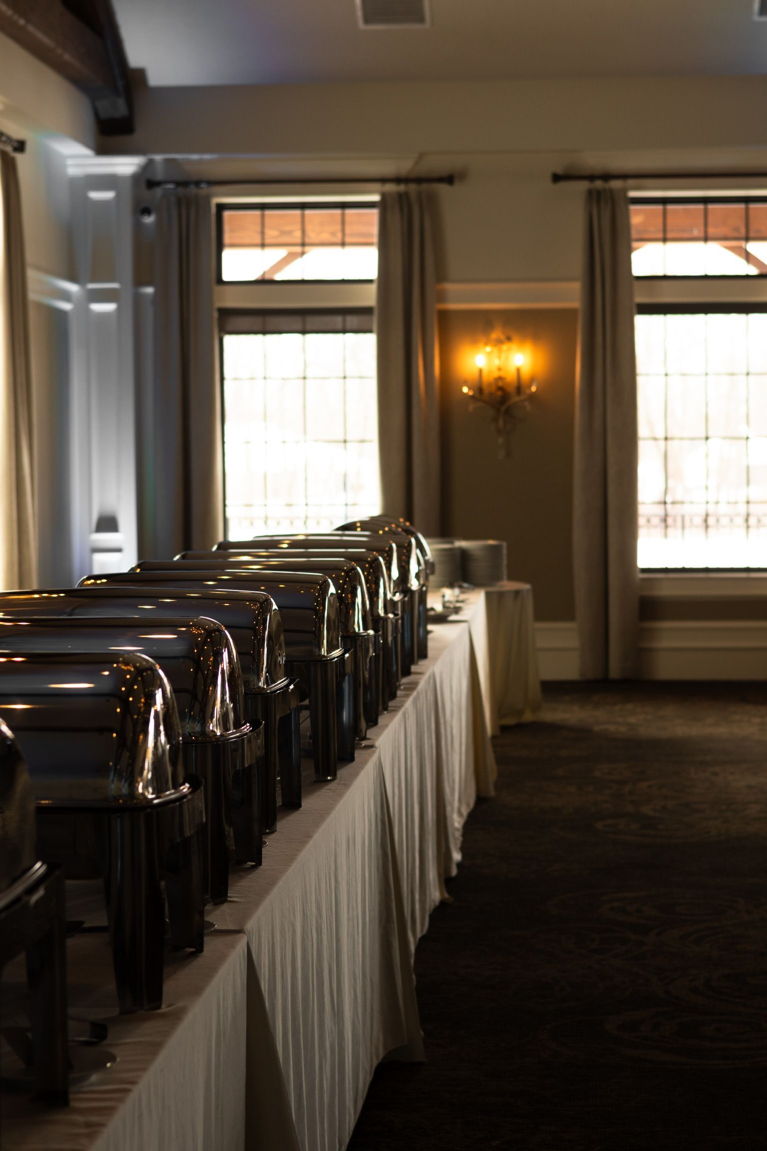 Buffet setup in a room with windows and sconces. Silver chafing dishes on a white tablecloth.