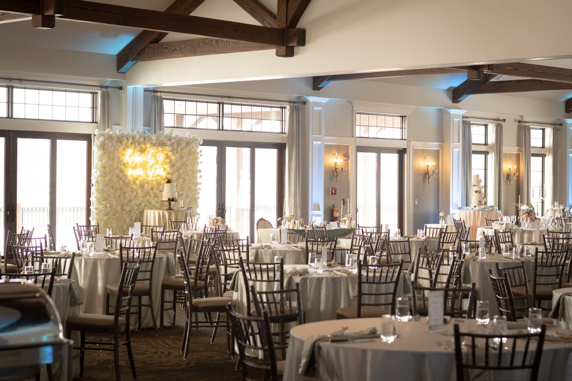 Elegant reception hall set for a wedding; round tables with white linens, brown chairs, floral backdrop, cake.